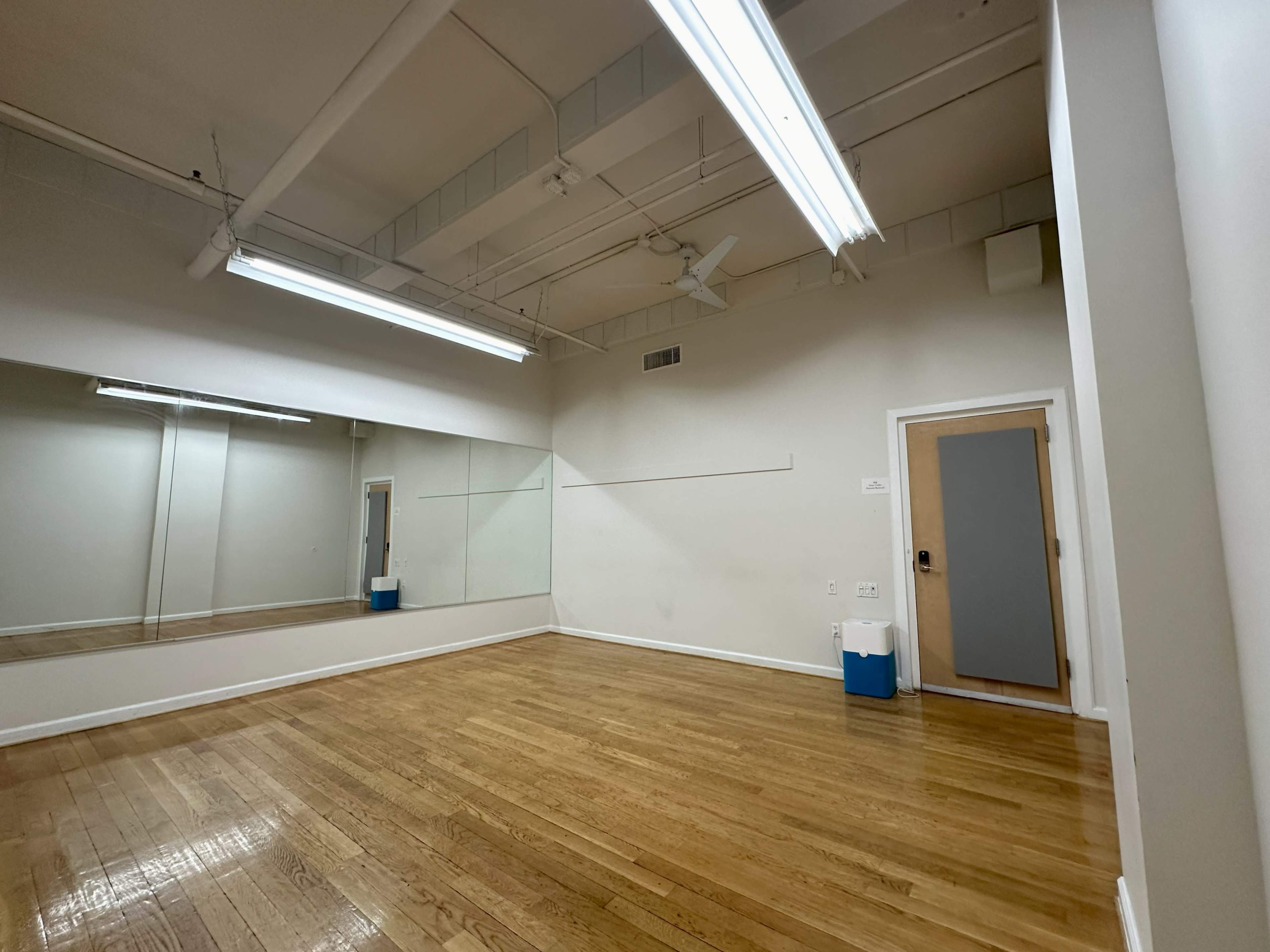 The image shows a spacious, empty room with wooden floors, a large mirror on one wall, and bright overhead lights.