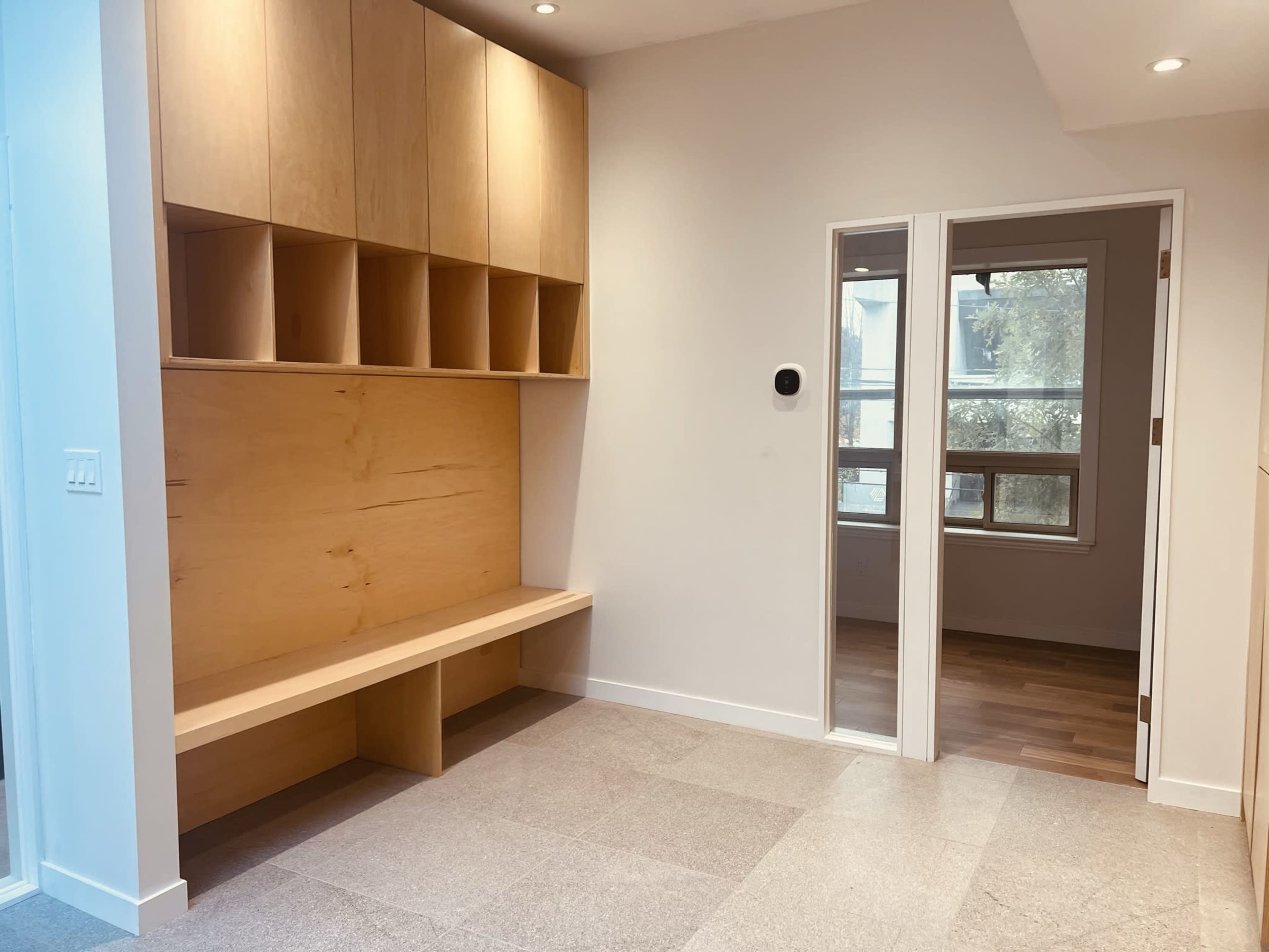 A modern, minimalist entryway with a wooden bench and shelving on one side, leading to a door opening into another room.