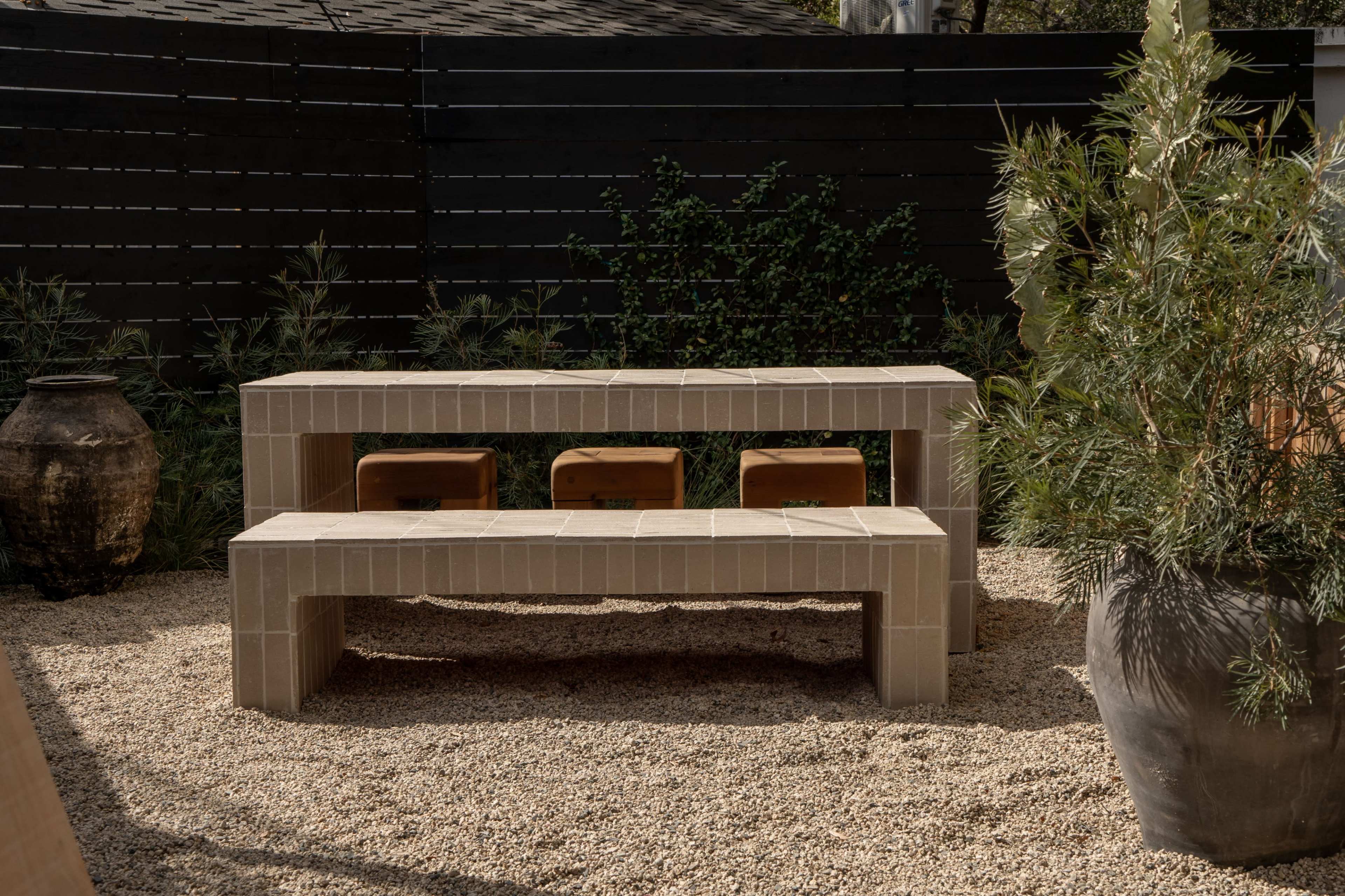 The image shows a stone bench and table set in a gravel garden surrounded by potted plants and a dark wooden fence.
