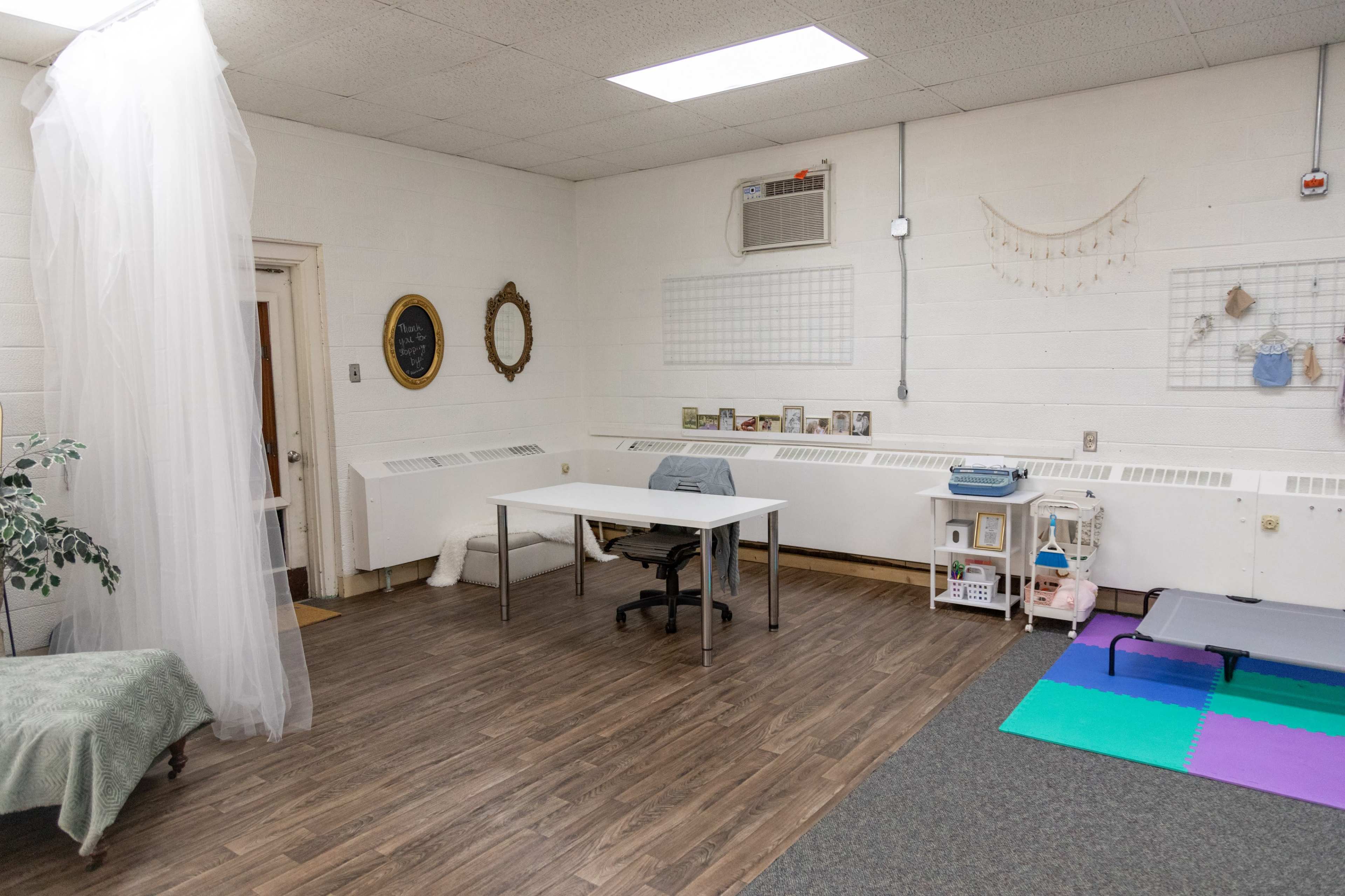 The image shows a sparsely furnished room with a desk and chair, a couch, wall decorations, and a soft floor mat.