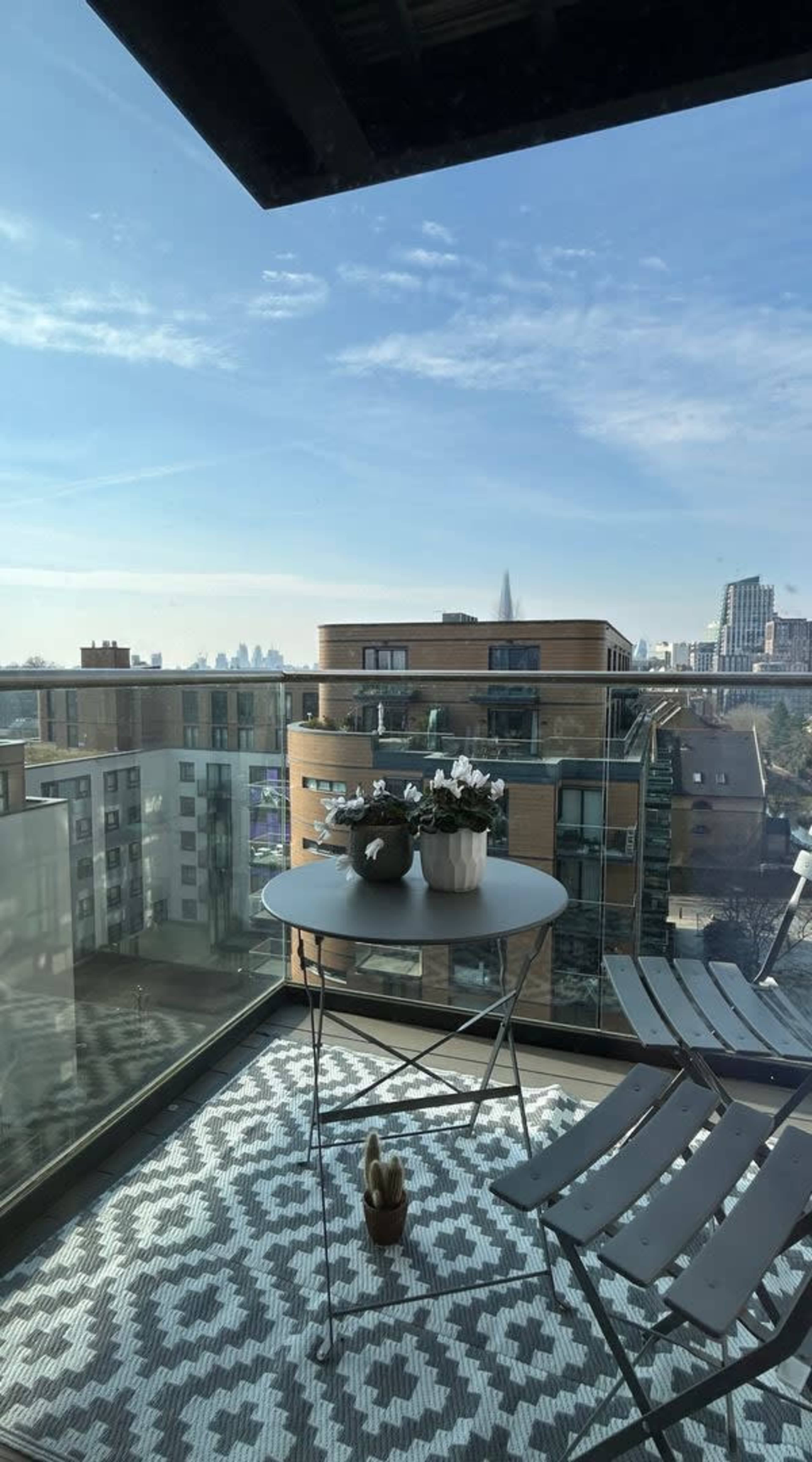 A minimalistic balcony features a round table with potted plants and a set of folding chairs, overlooking a cityscape under a clear sky.
