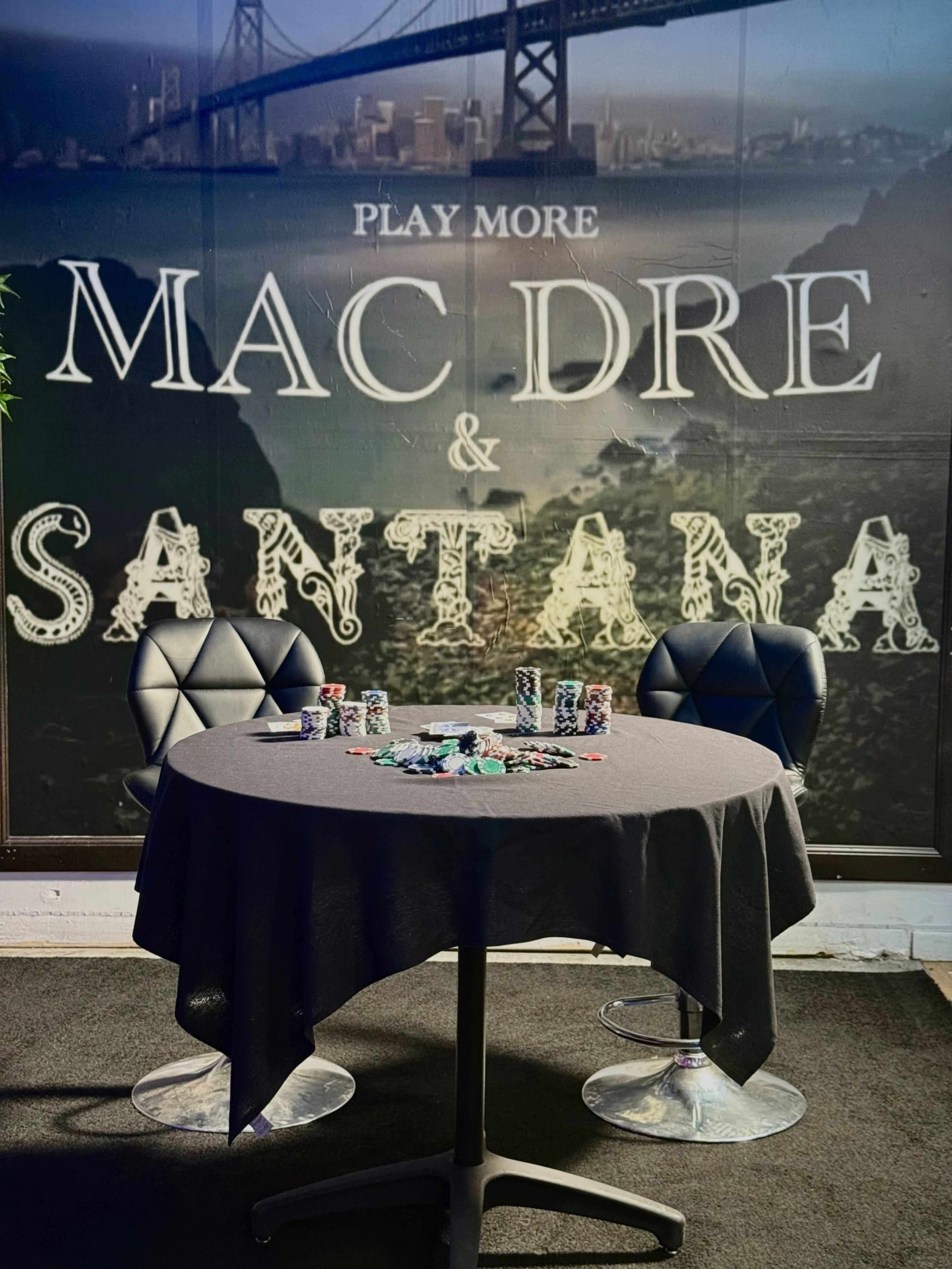 A table with a black tablecloth is set with colorful cups and chips, positioned in front of a mural featuring "MAC DRE" and "SANTANA" along with a view of a bridge.
