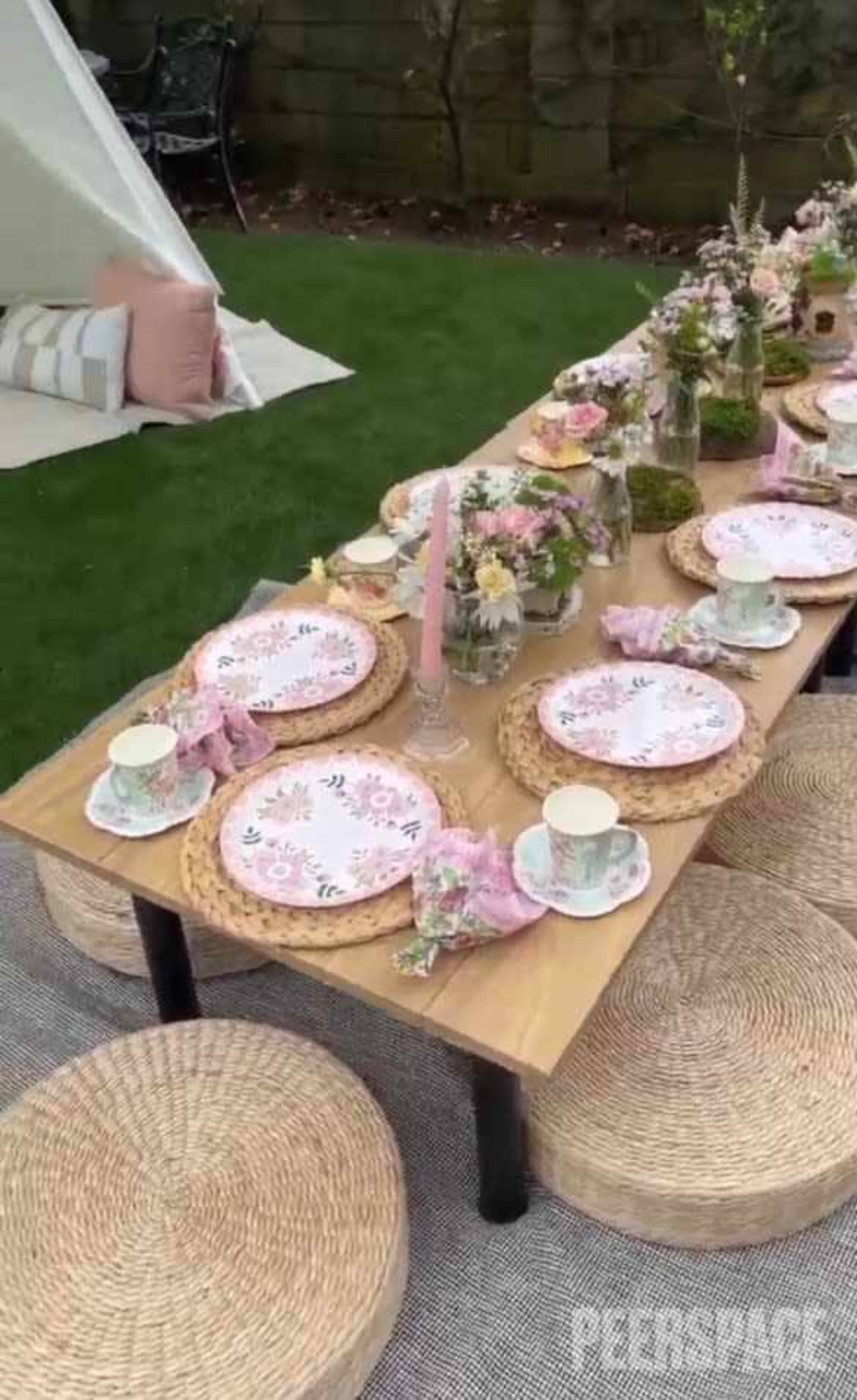 A long wooden table is set outdoors with floral-patterned plates, cups, and decorative elements, surrounded by woven seating.