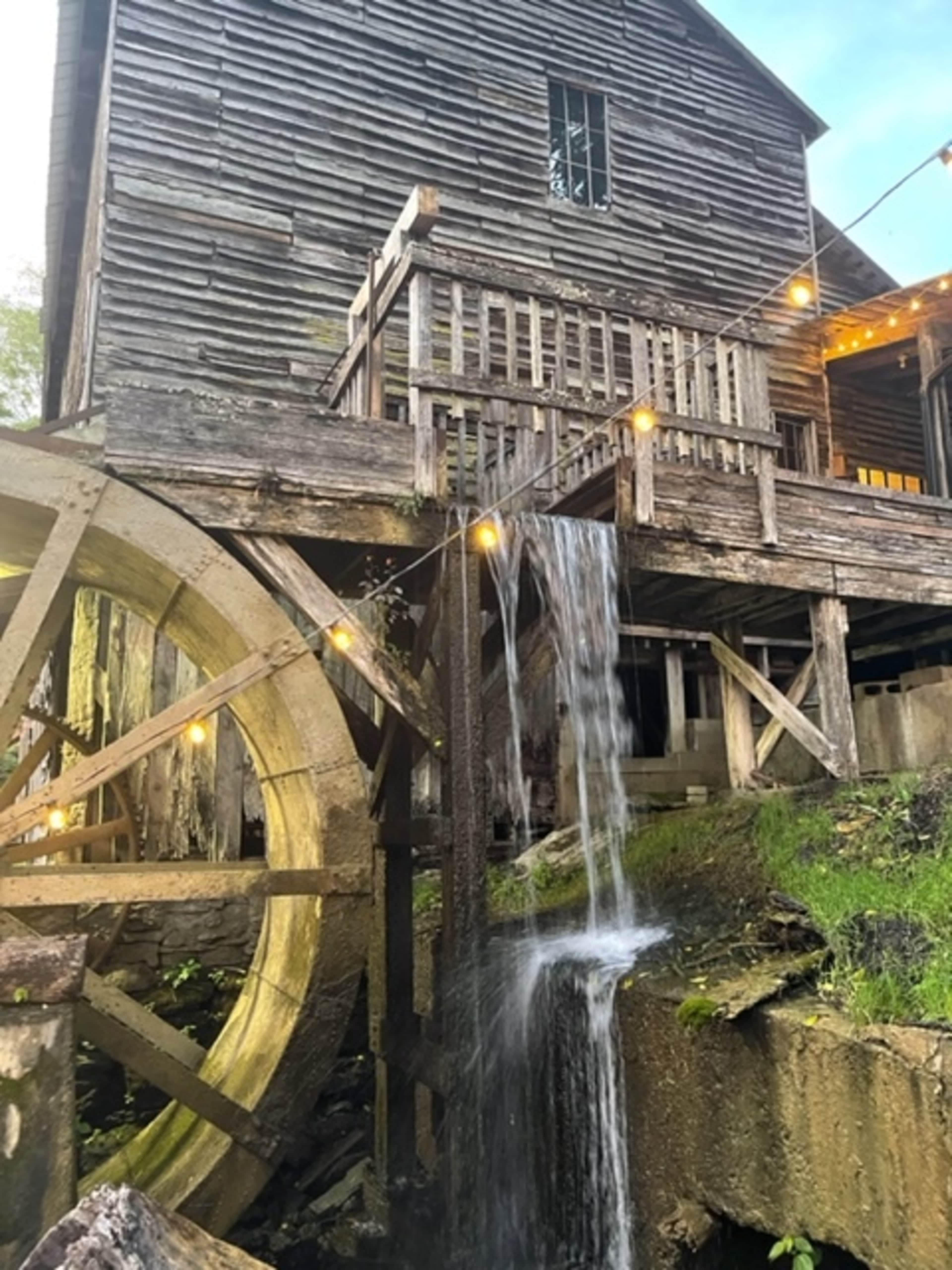 A wooden building with a waterwheel, where water cascades down over the wheel and onto the ground below, illuminated by string lights above.