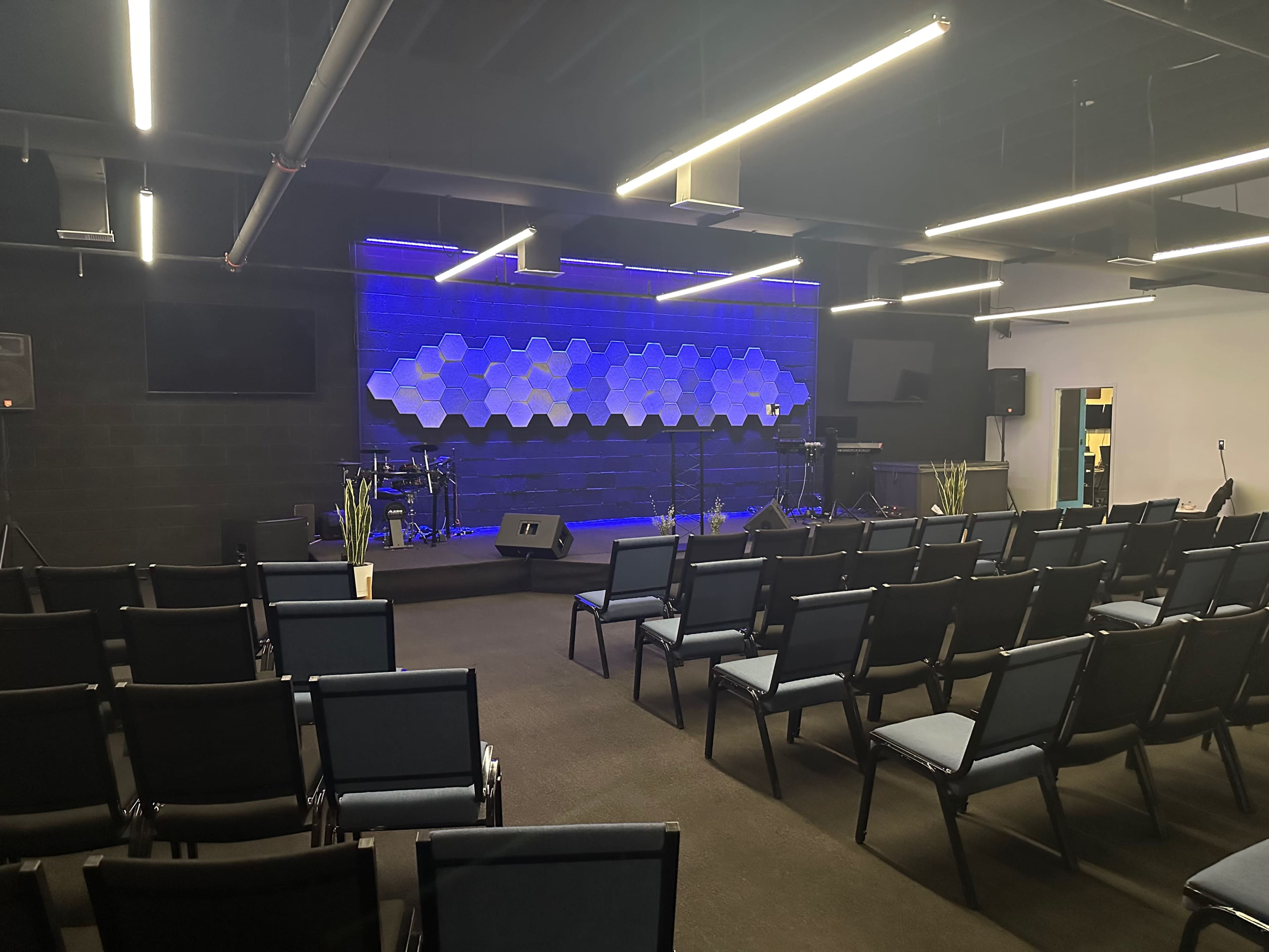 A modern interior of a performance space with rows of chairs facing a stage featuring a hexagonal backdrop, musical instruments, and lighting.