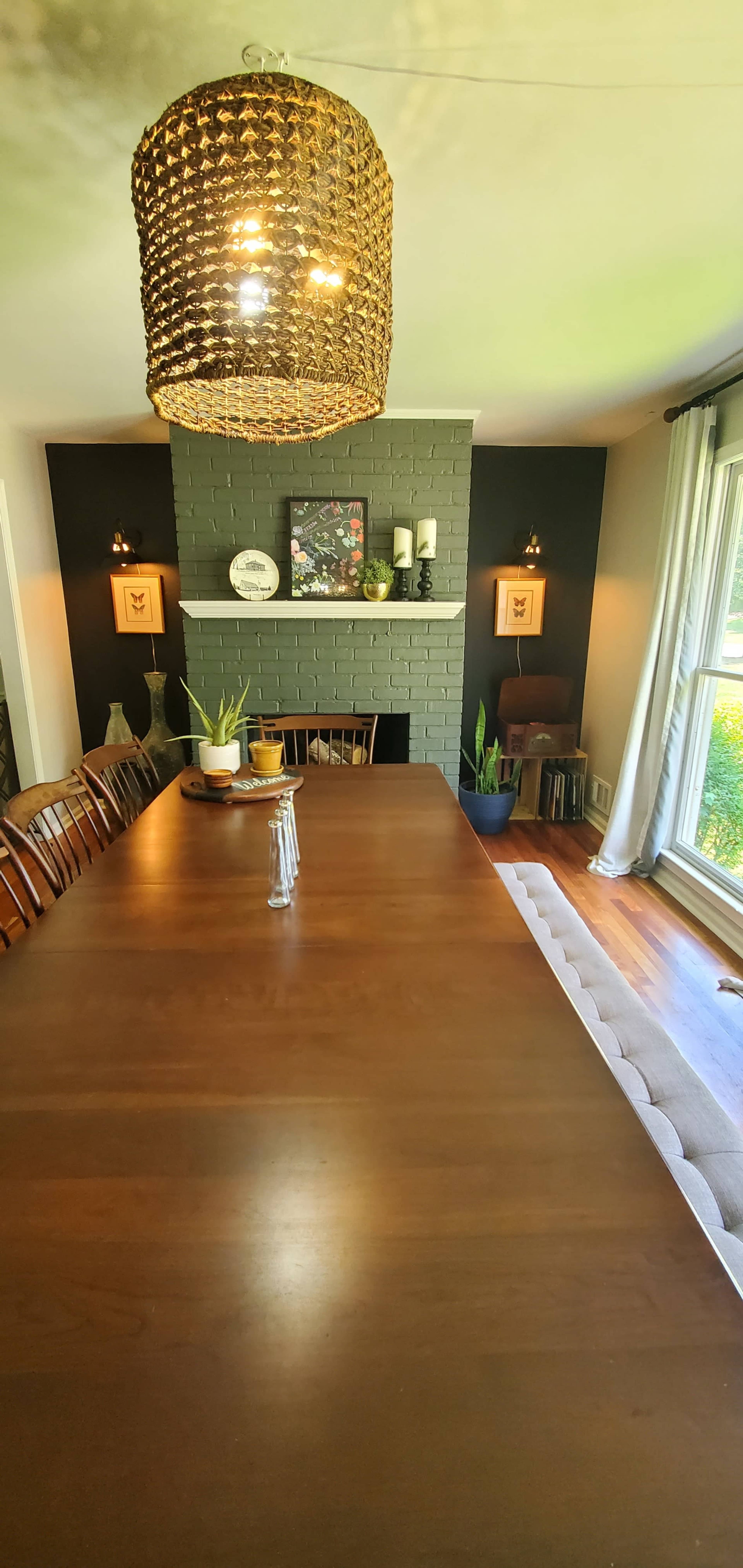 A long wooden dining table is set in a well-lit room featuring a fireplace with decorative elements and large windows overlooking greenery.