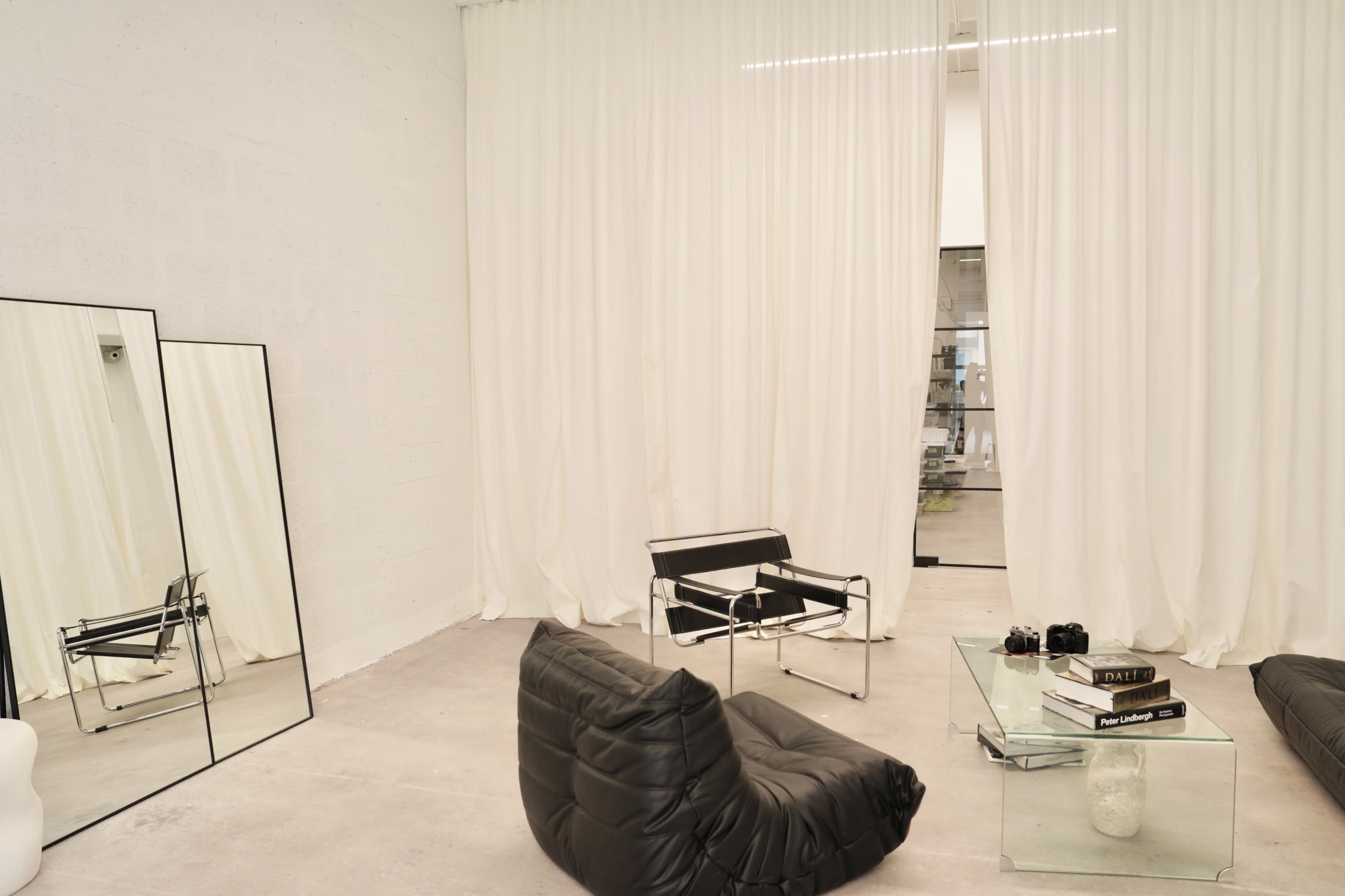 The scene features a minimalist interior with a black contemporary chair, a mirrored reflection, a glass coffee table with books and a camera, and white curtains hanging in the background.