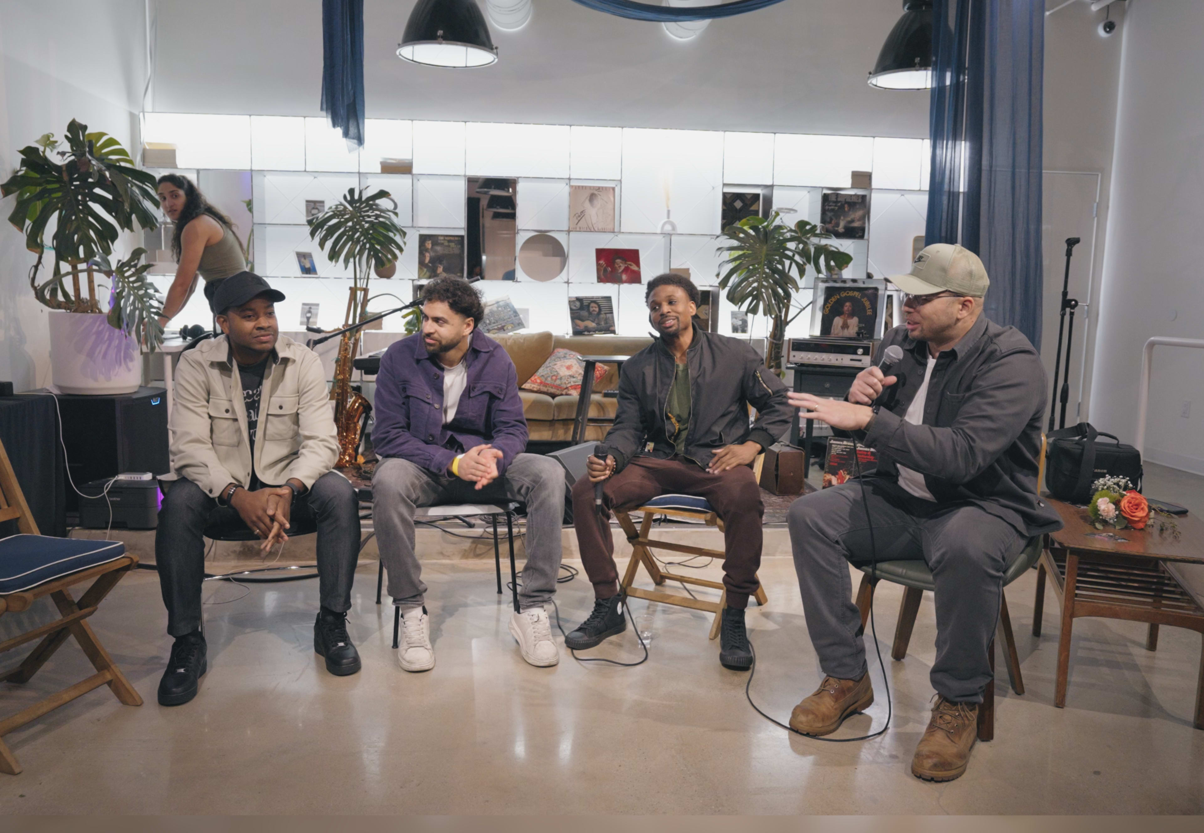A group of four men sit on chairs in a modern, decorated space, engaging in conversation while a woman is visible in the background.