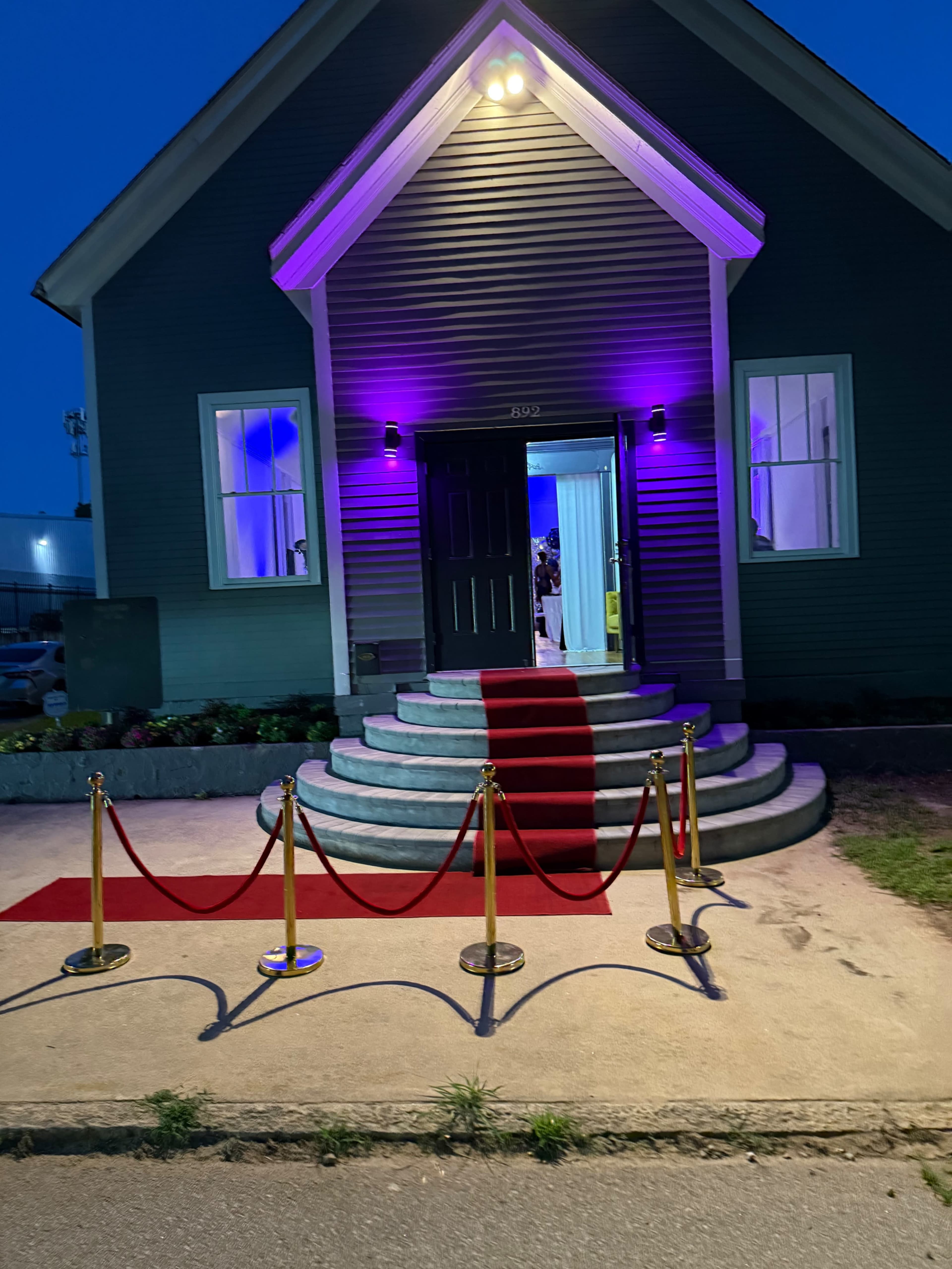 A building with a dark green exterior is illuminated by purple lights, featuring a red carpet leading up to its entrance, which is flanked by stanchions with ropes.