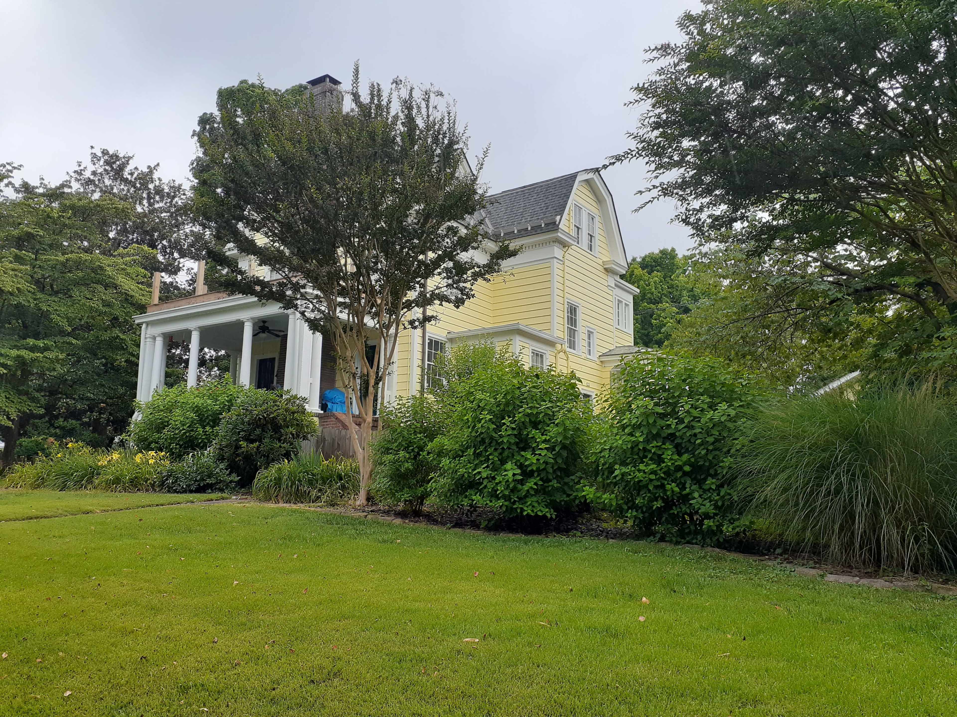 A two-story yellow house with a porch is surrounded by shrubs and trees in a green lawn.