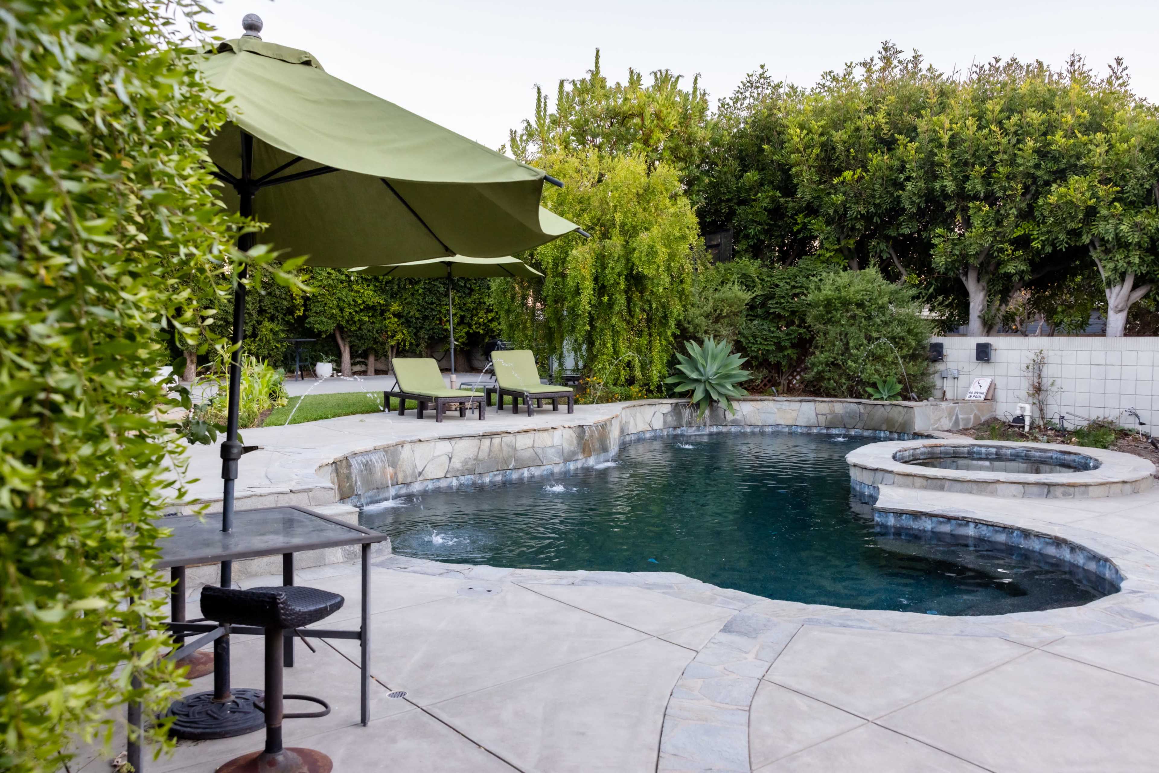 A serene backyard features a pebble-finished pool with a spa, surrounded by lounge chairs, a table, and lush greenery.