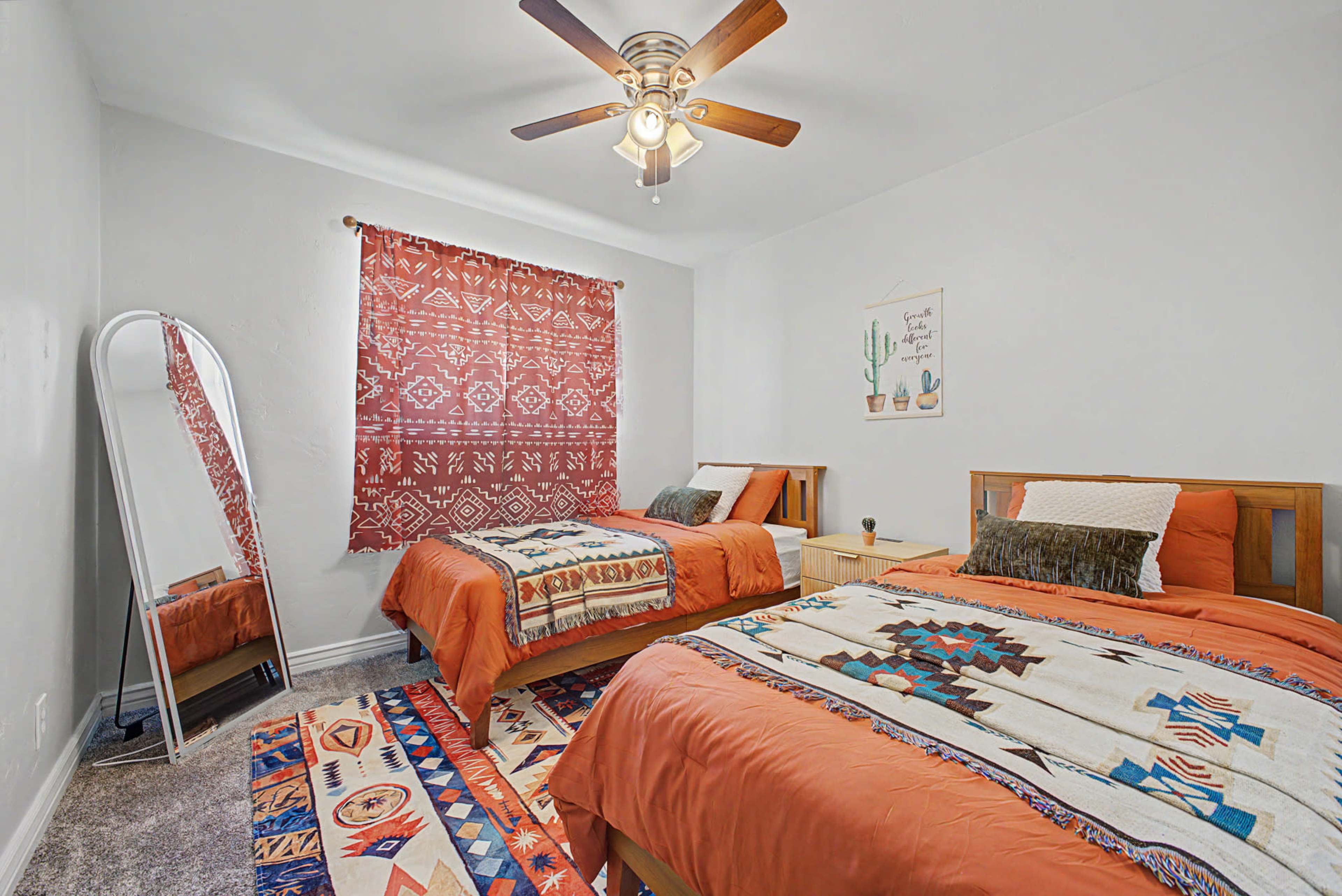 The image shows a well-decorated bedroom featuring two beds with orange bedding, a patterned rug, a mirror, and a window covered with a red patterned curtain.