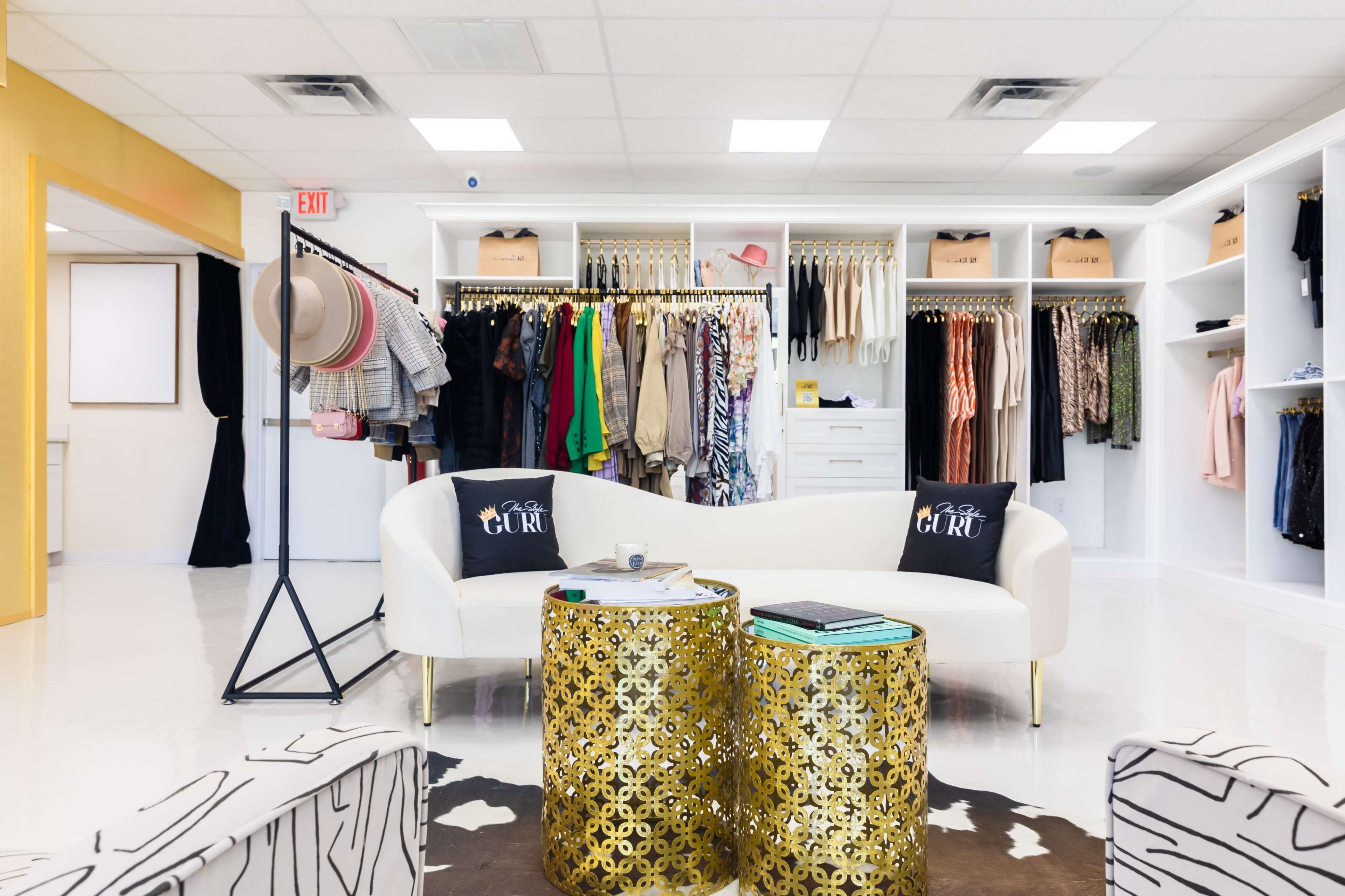 A retail boutique interior featuring a white sofa with decorative pillows, two golden side tables, and clothing racks displaying various garments.