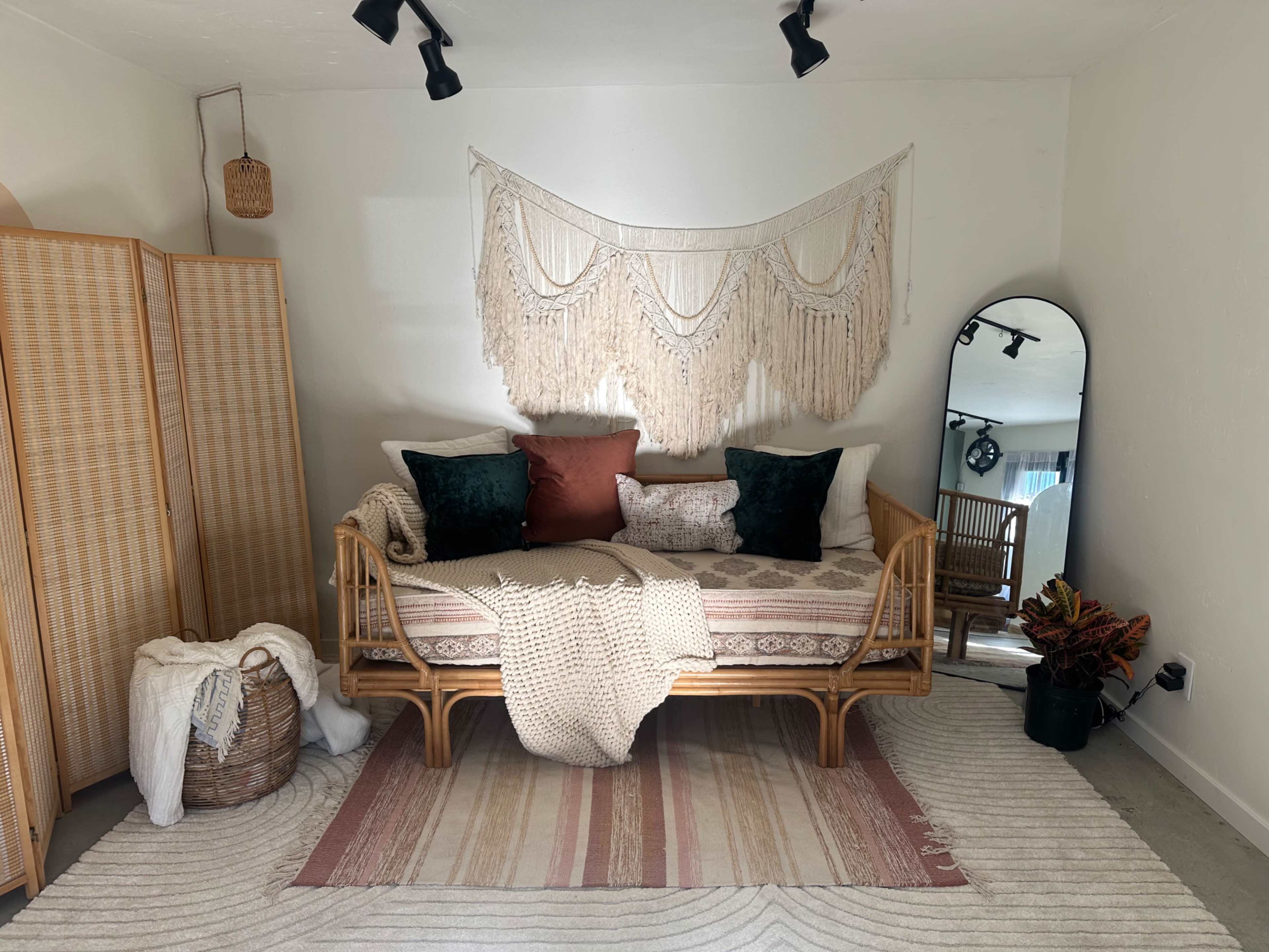 The image shows a cozy corner featuring a daybed adorned with pillows, a large mirror, a woven basket, and a decorative macramé wall hanging.