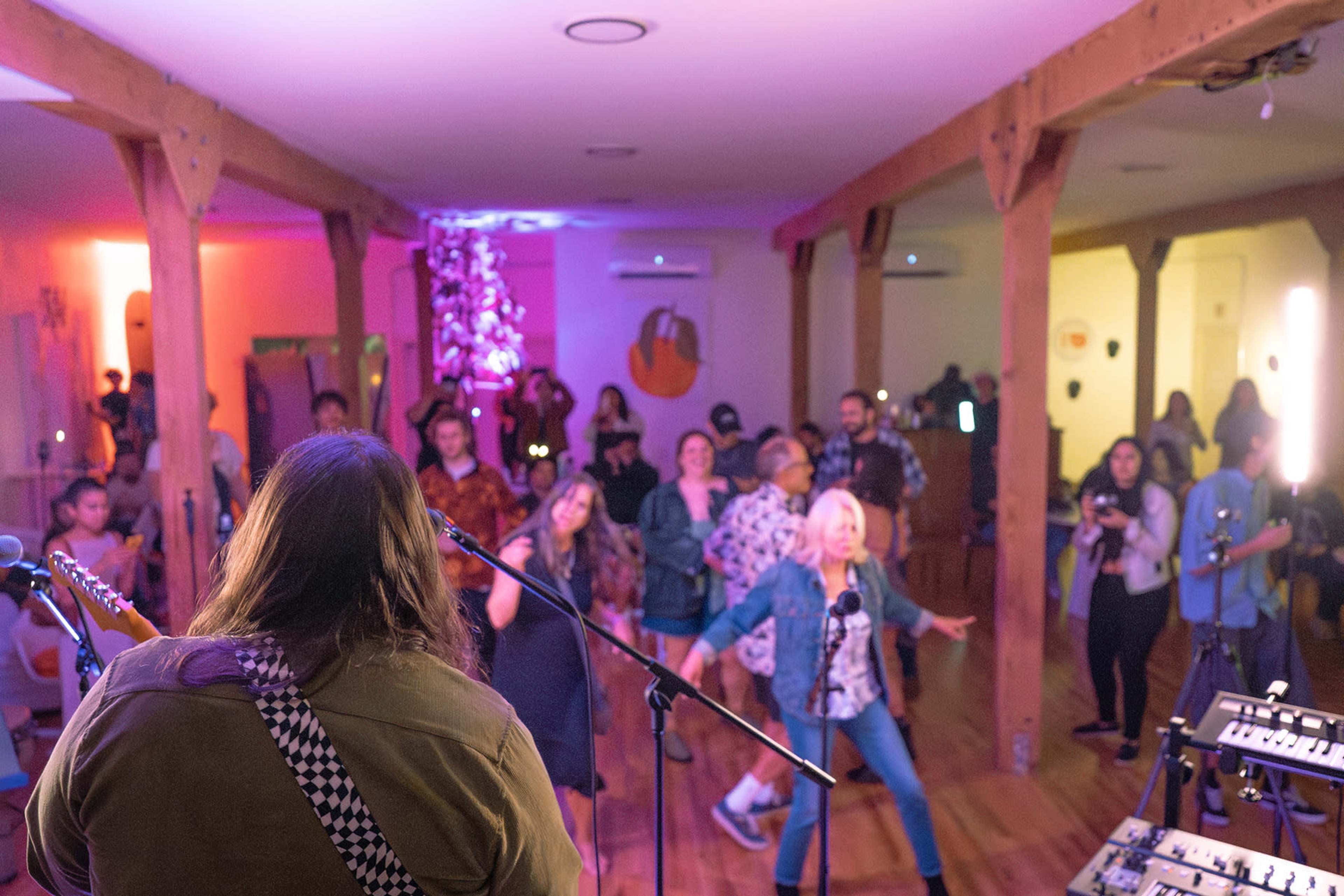 A crowd of people enjoys a live music performance in a warmly lit room with wooden beams and colorful decor.