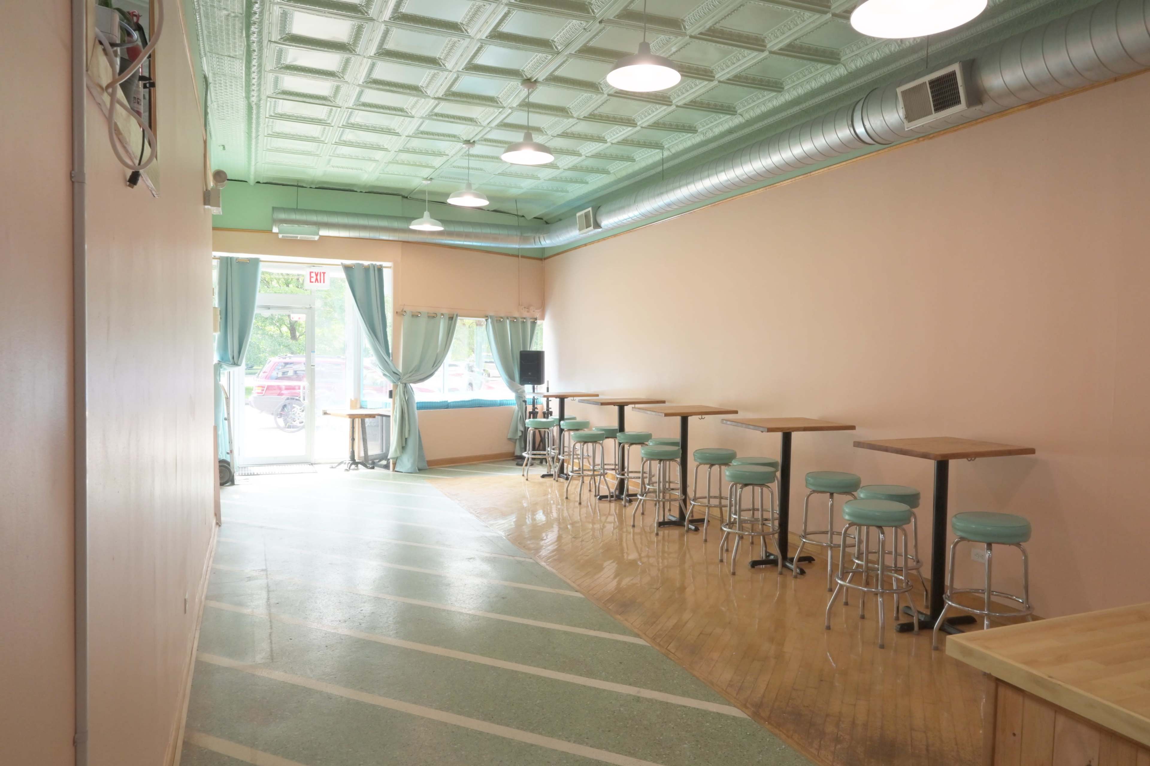 A narrow, vacant café interior with a light pink wall, metal stools along the side, and large windows allowing natural light to enter.
