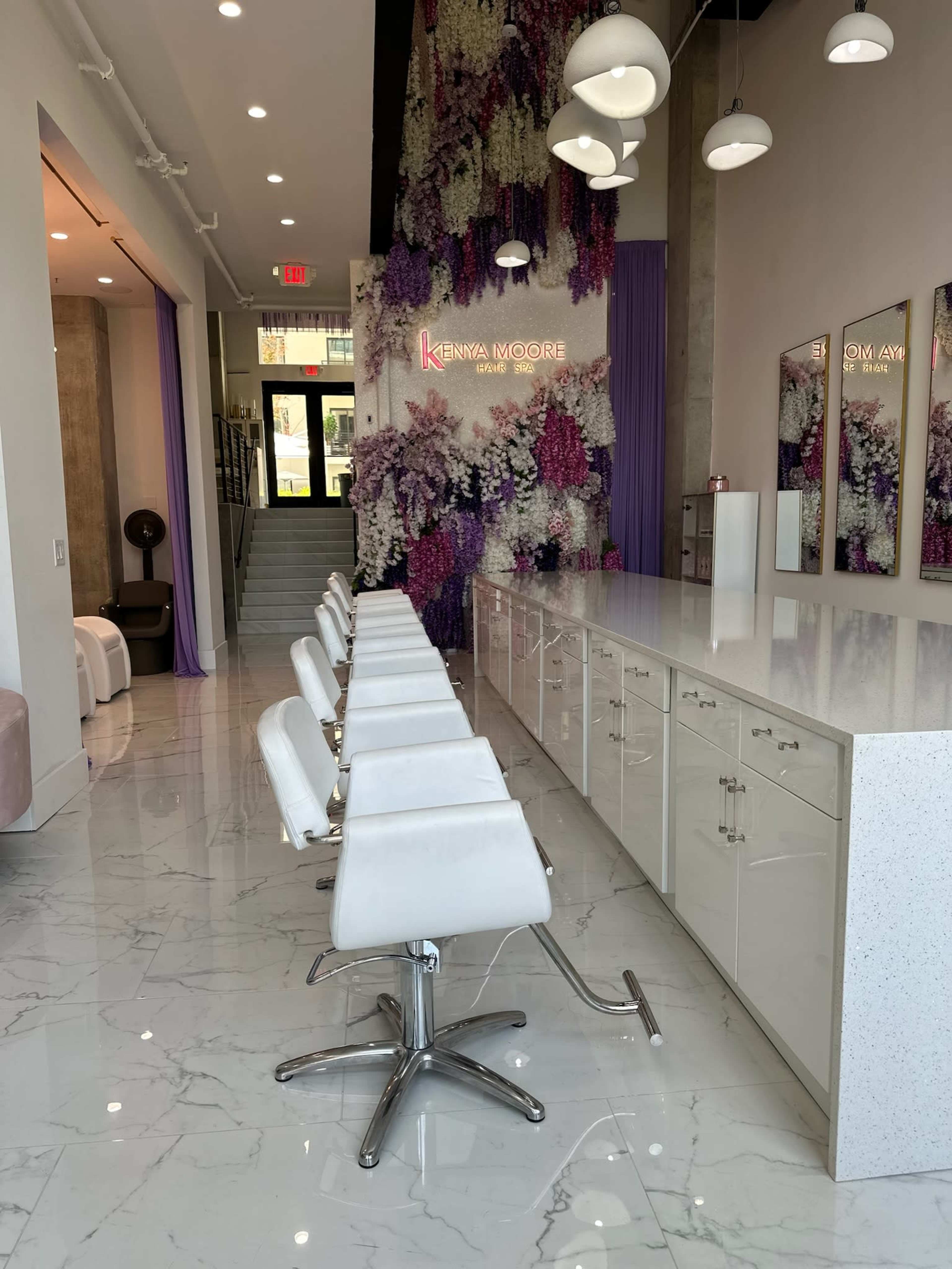 The image shows a modern salon interior with white styling chairs lined up in front of a white countertop, and a wall adorned with purple and white flowers.