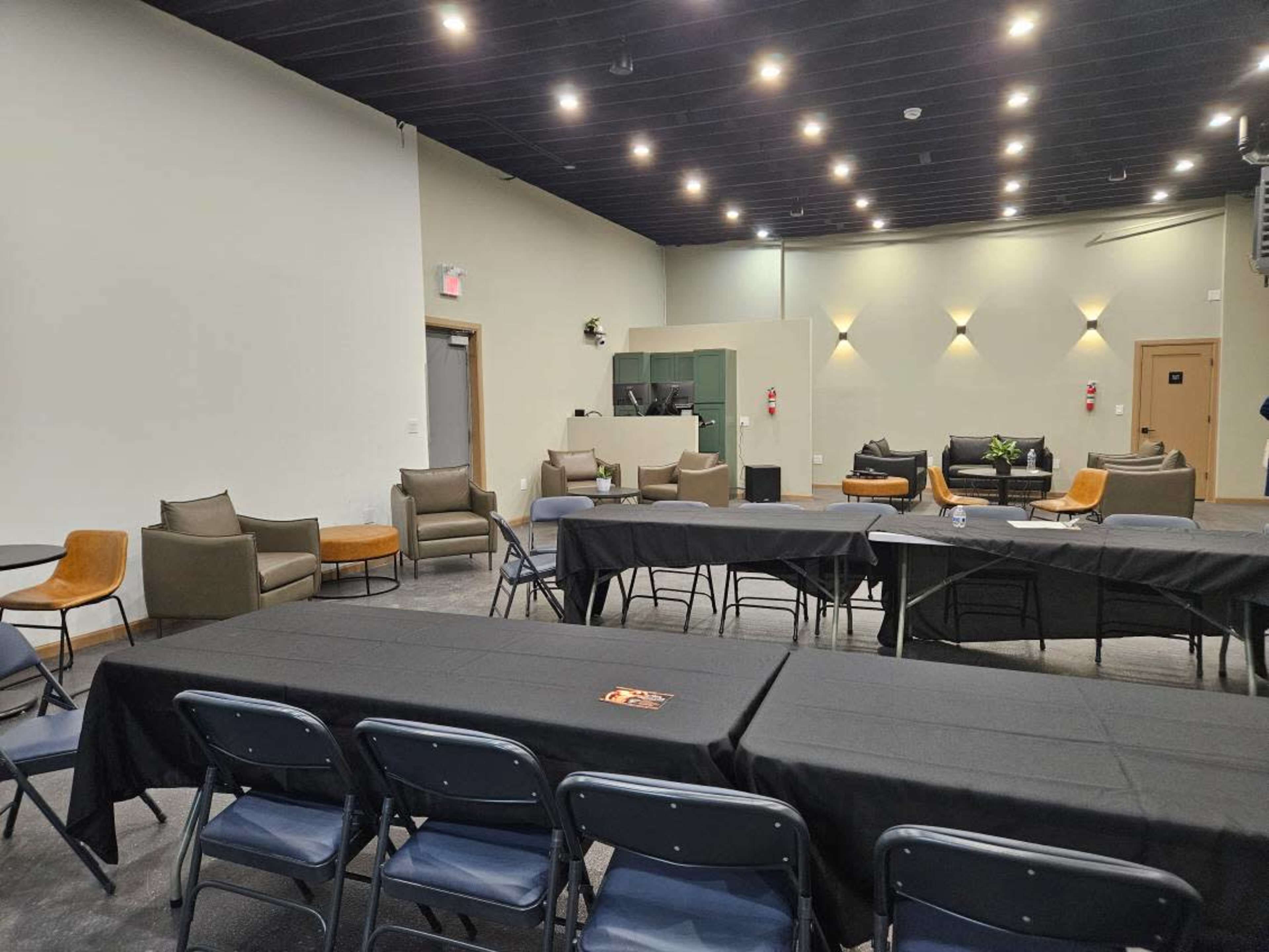The image shows a modern, empty meeting space with several tables covered in black cloth, chairs arranged around them, and a lounge area in the background.
