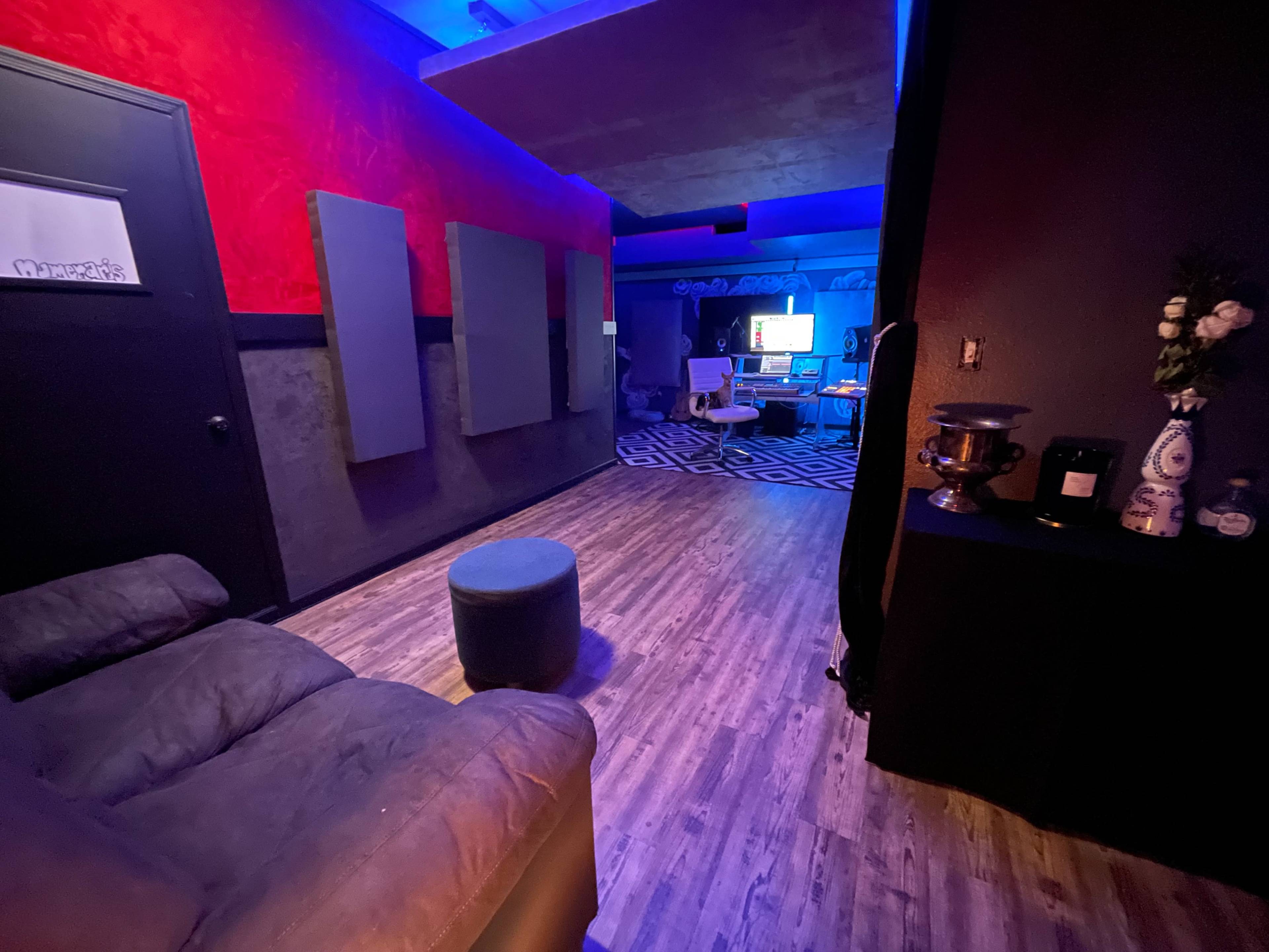 A dimly lit room features a gray couch, a circular ottoman, and a workspace in the background with colorful lighting and musical equipment.