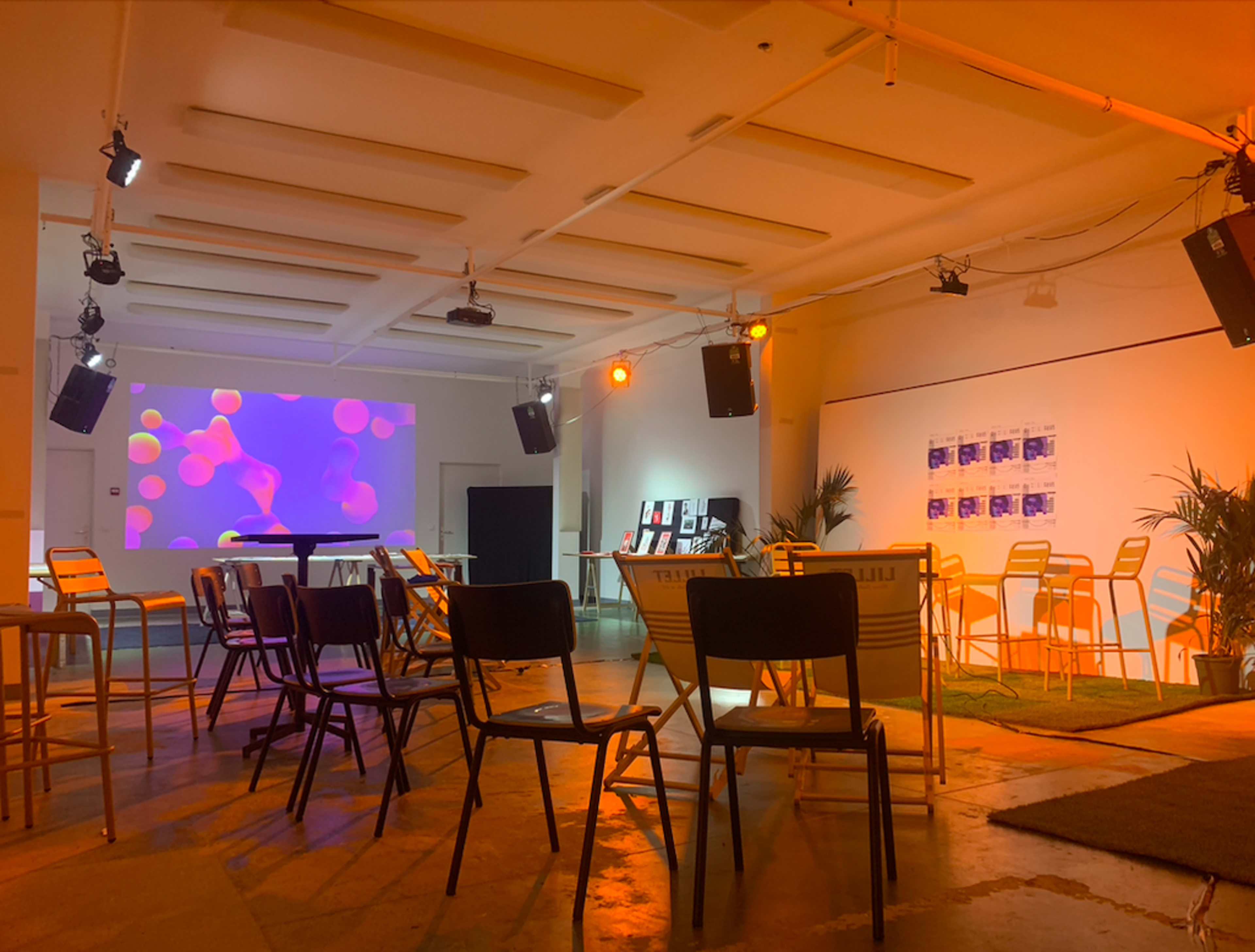 A spacious room with chairs arranged in rows facing a large screen displaying colorful visuals, complemented by a few potted plants and speakers mounted on the walls.