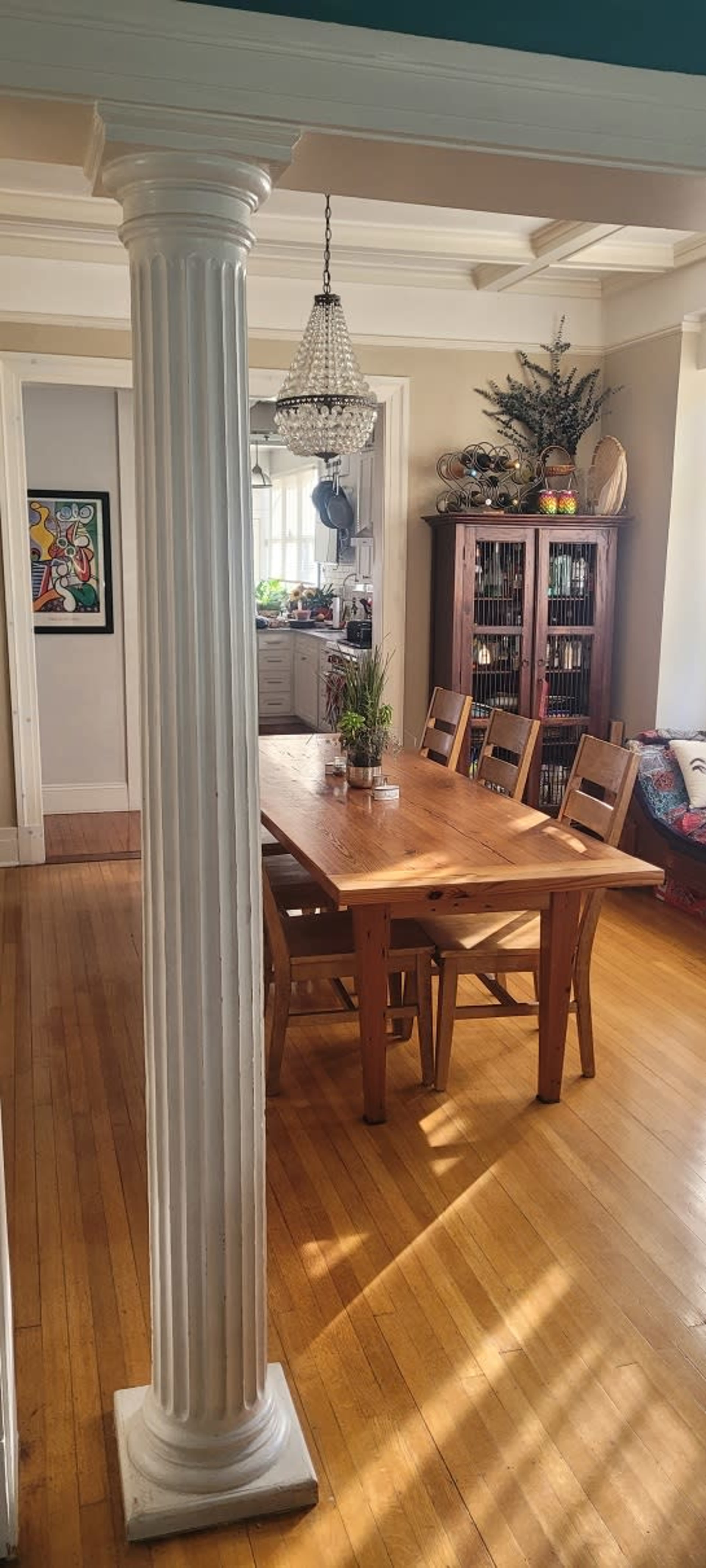 The image shows a dining room with a wooden table and chairs, a column, and a glass chandelier, illuminated by natural light.