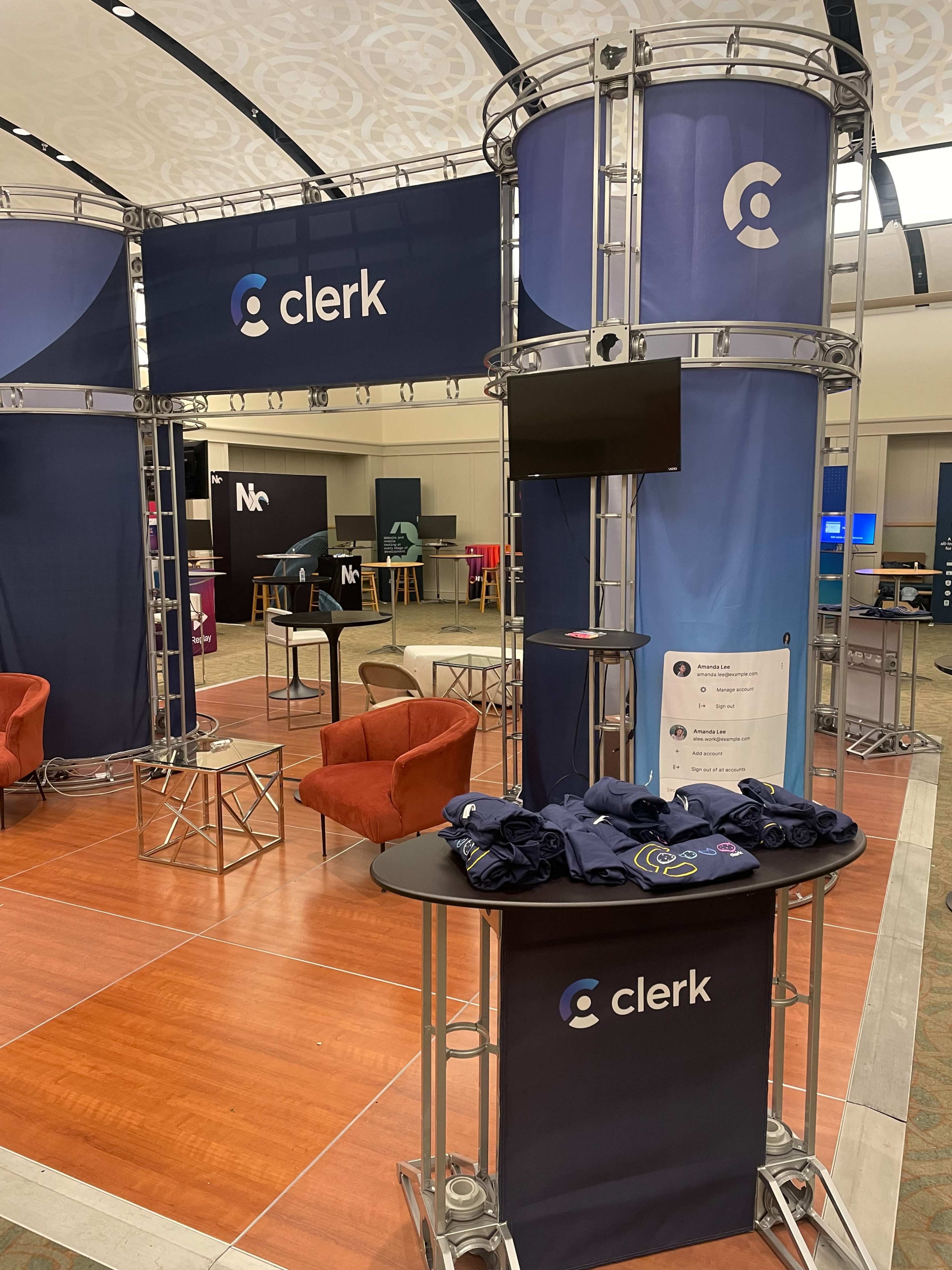 The image shows a display booth for a company called "clerk," featuring a curved structure, promotional materials, a television screen, and branded merchandise on a table in a convention center.