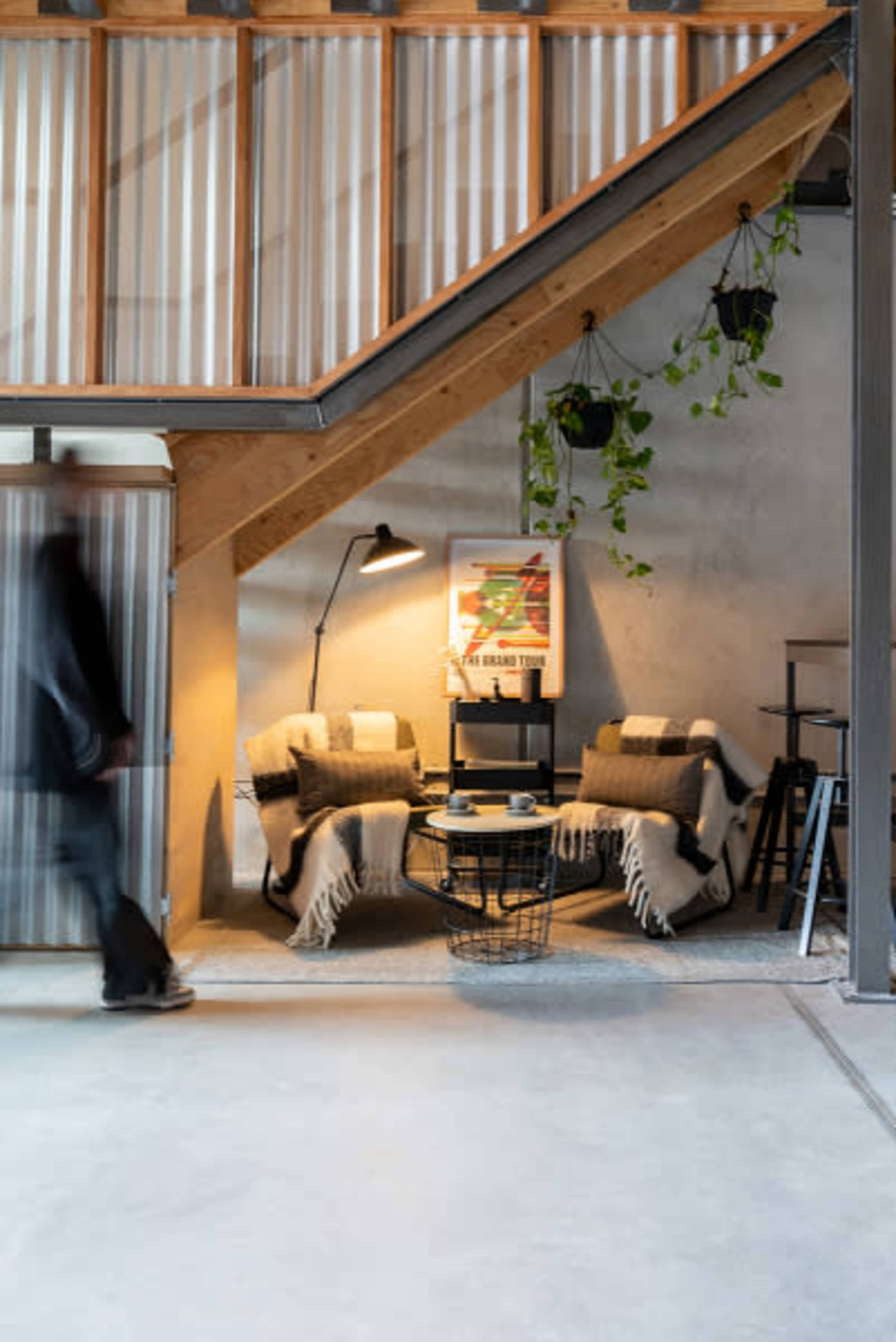 A person walks past a cozy seating area with two chairs, a small table, and decorative plants under a wooden staircase.
