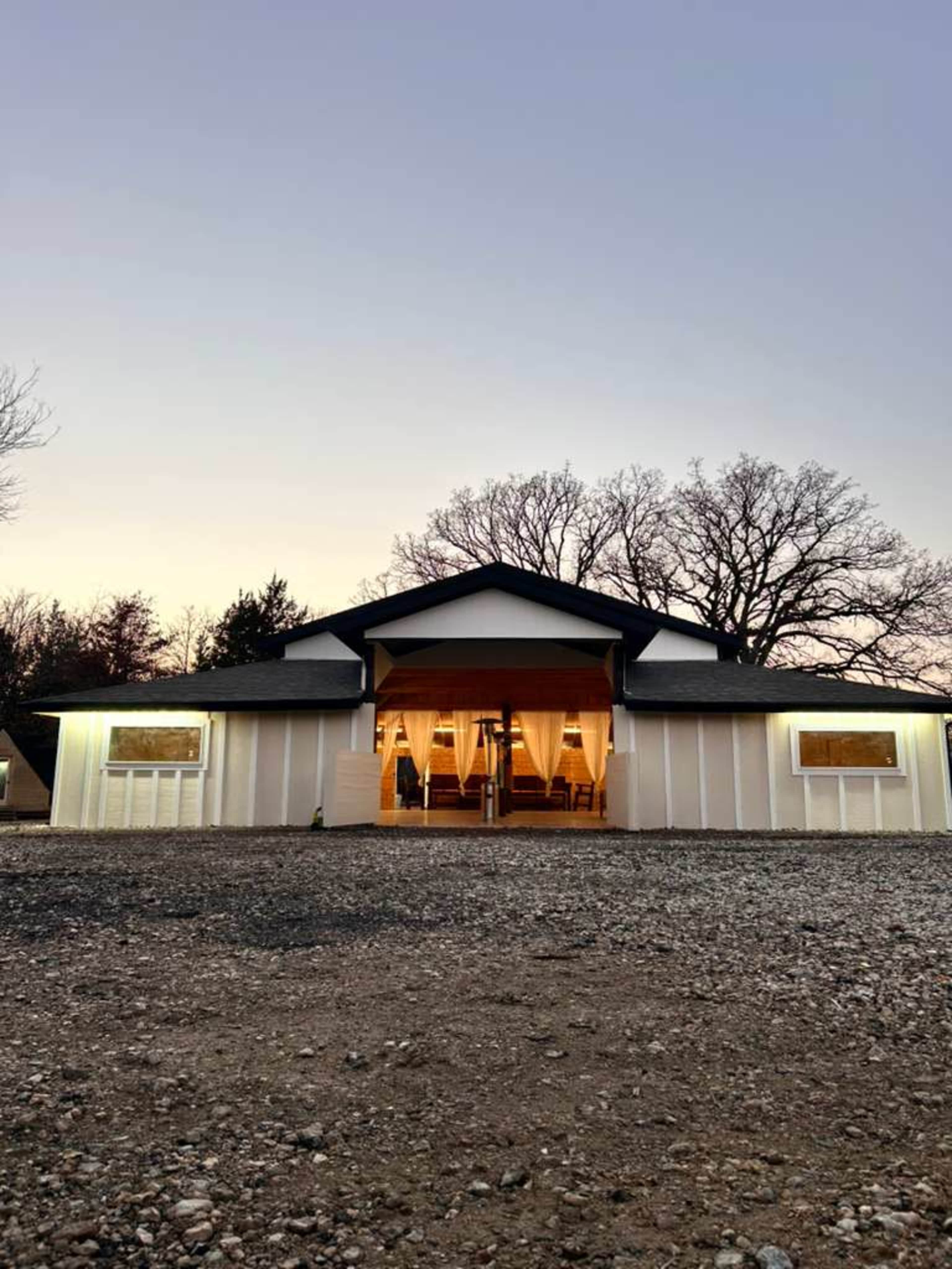 A rustic building with a gabled roof and large front windows is set against a twilight sky, surrounded by gravel.