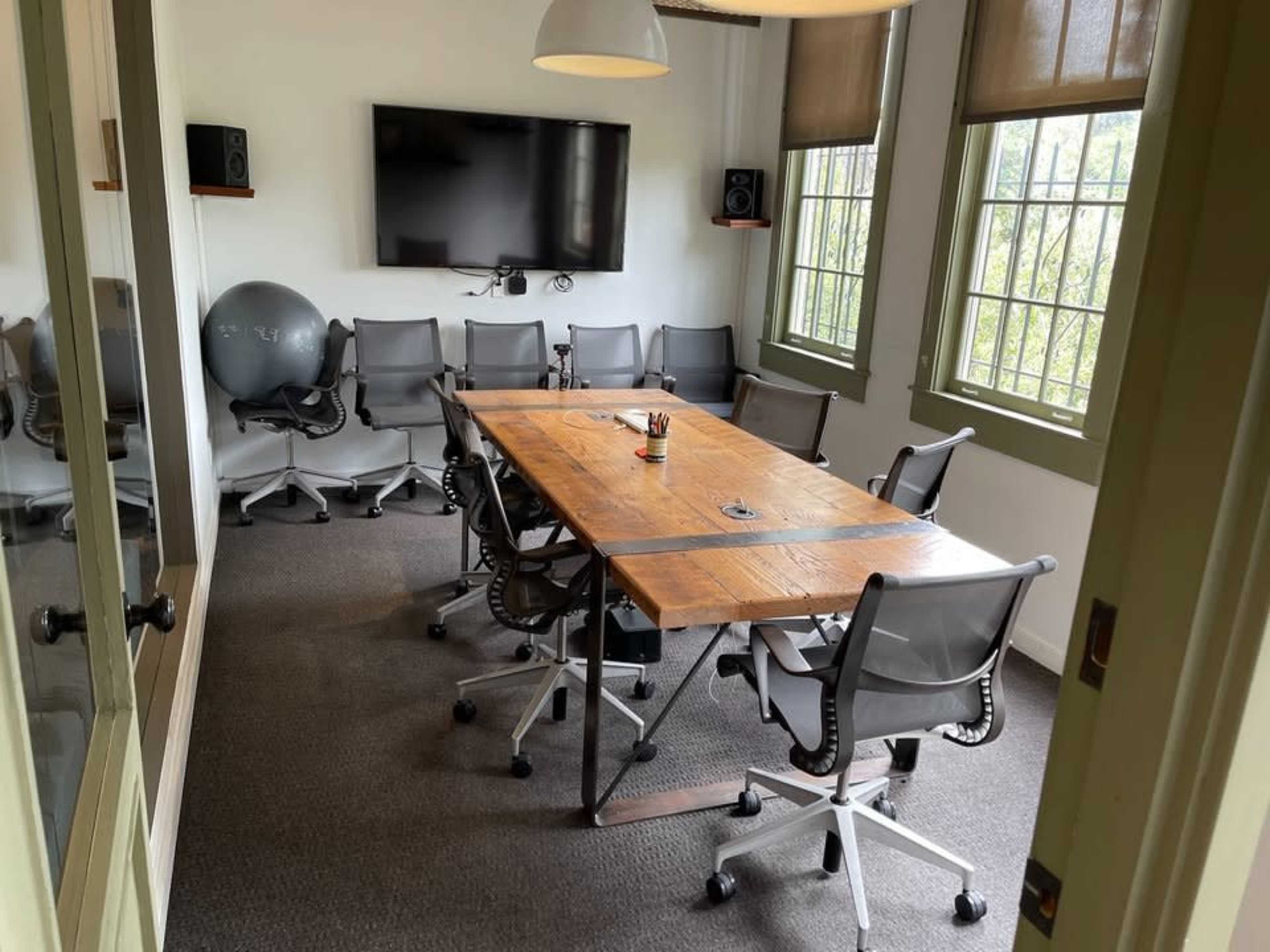 The image shows a conference room with a large wooden table surrounded by several ergonomic chairs, a large screen mounted on the wall, and a large exercise ball in one corner.