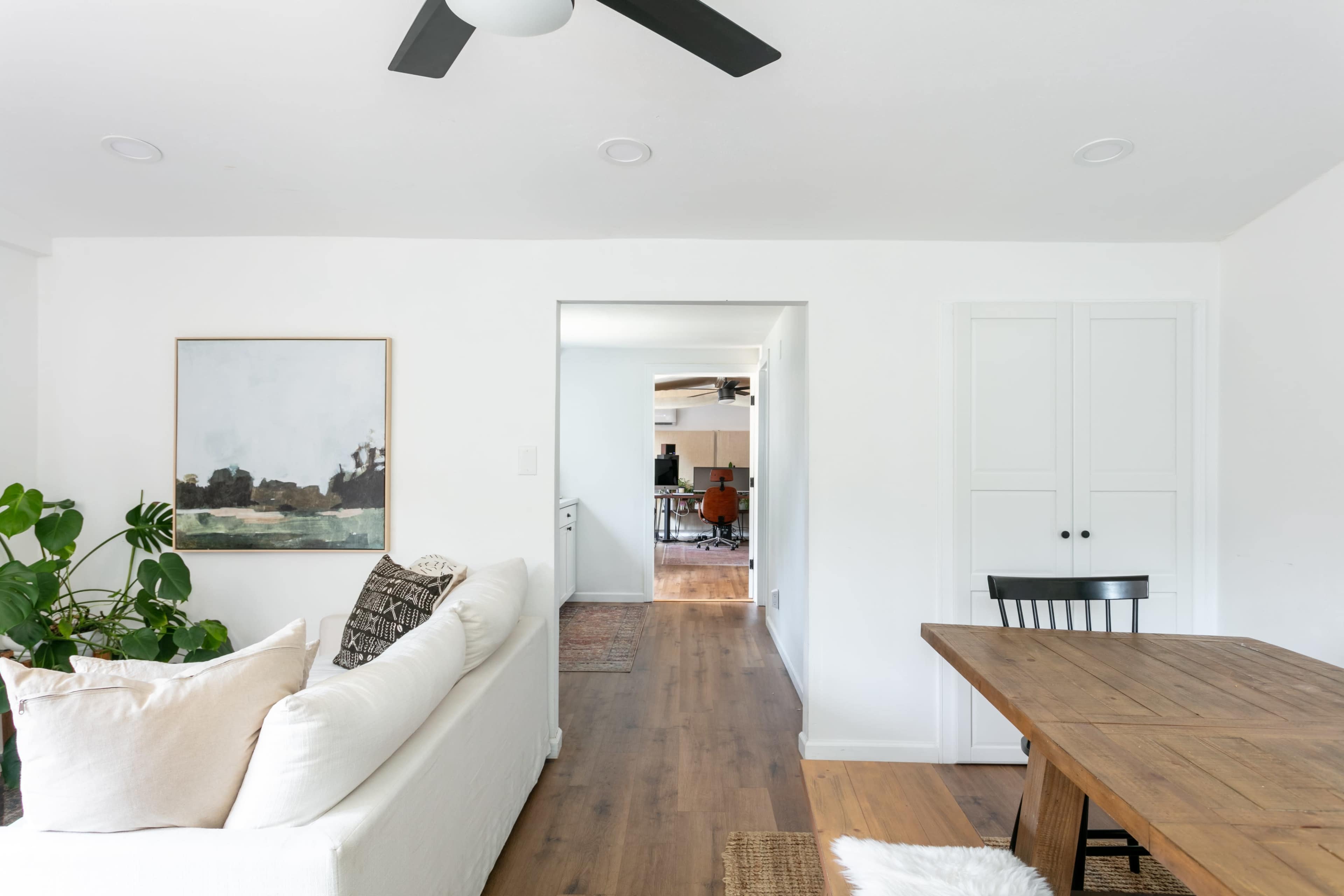 The image shows a bright, open living space with a couch, a dining table, and a doorway leading to another room.