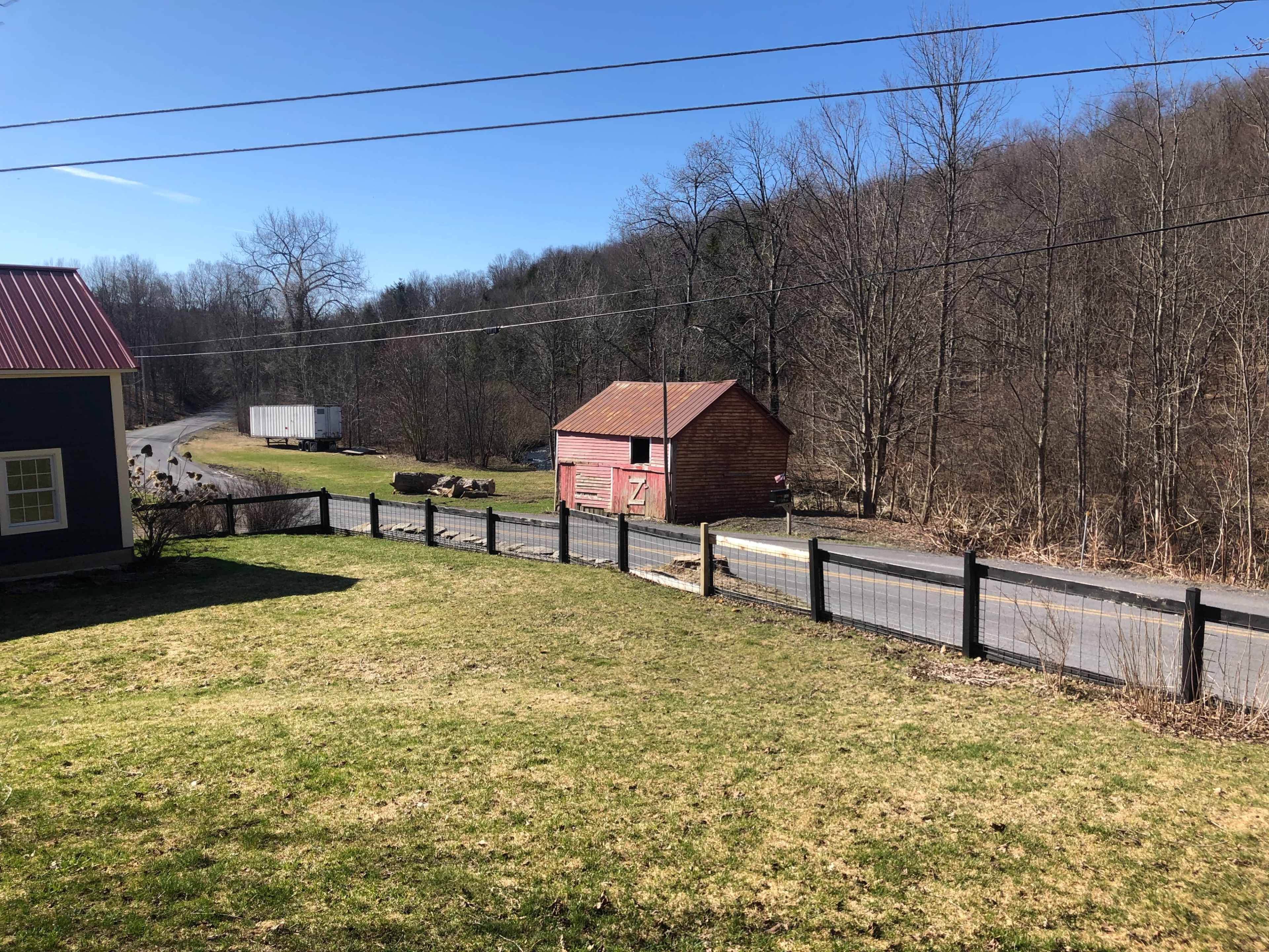 The image shows a grassy yard with a black fence, a winding road, a red barn, and trees in the background under a clear blue sky.