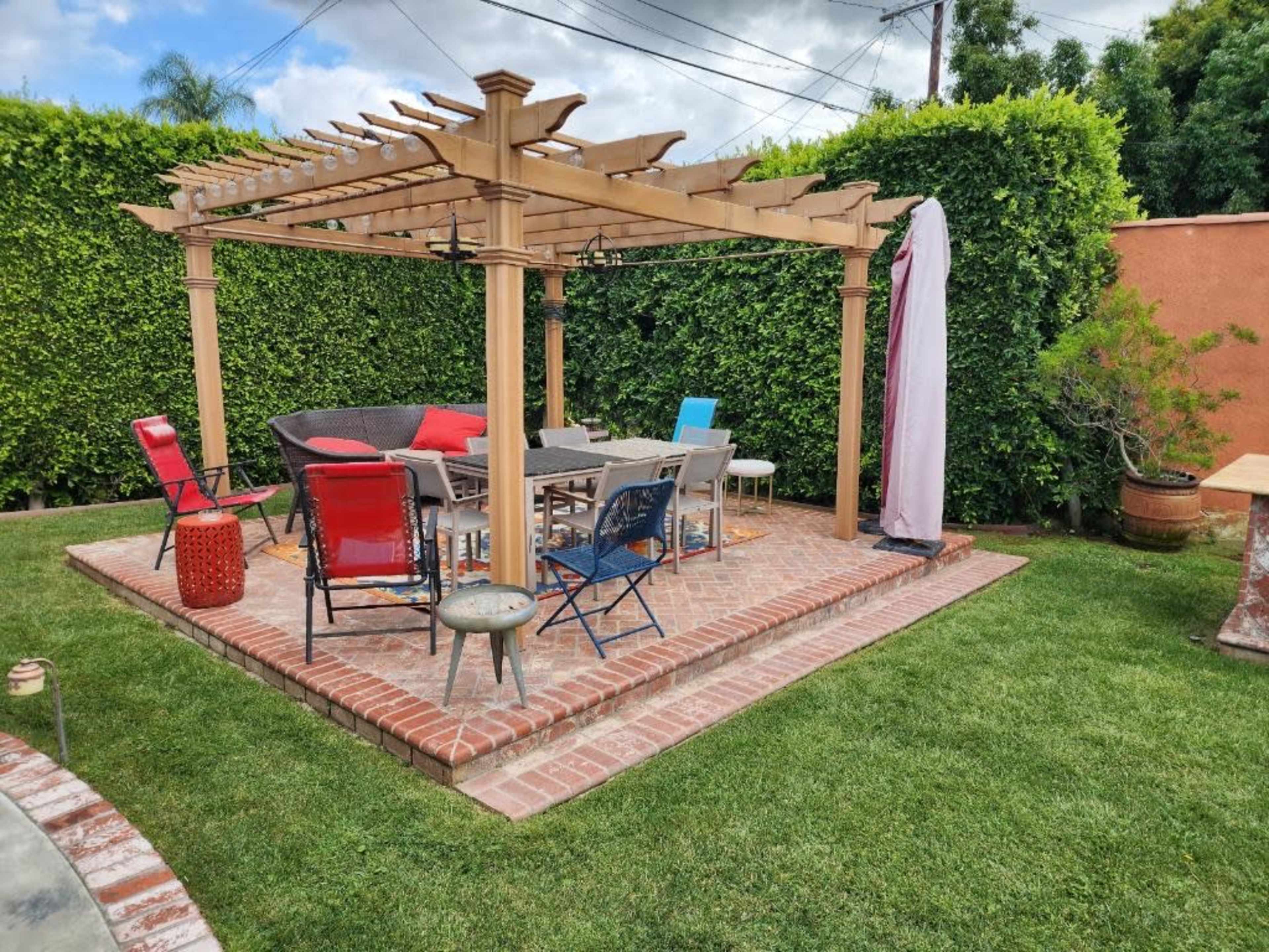 A outdoor patio features a wooden pergola with seating arrangements, surrounded by green hedges and brick pavement.