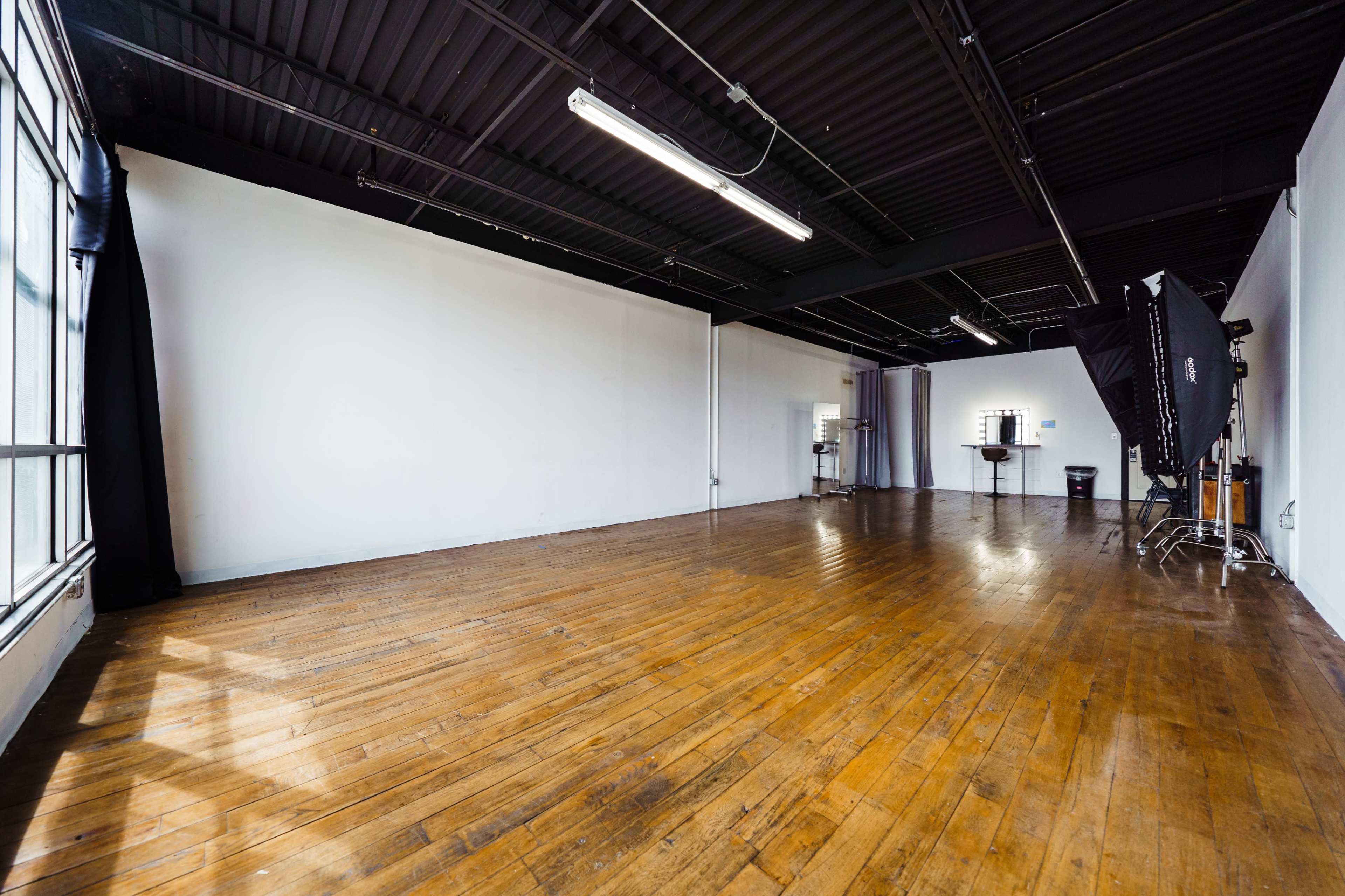 A spacious, empty studio features wooden flooring and large windows, with minimal furnishings and a photography backdrop in one corner.