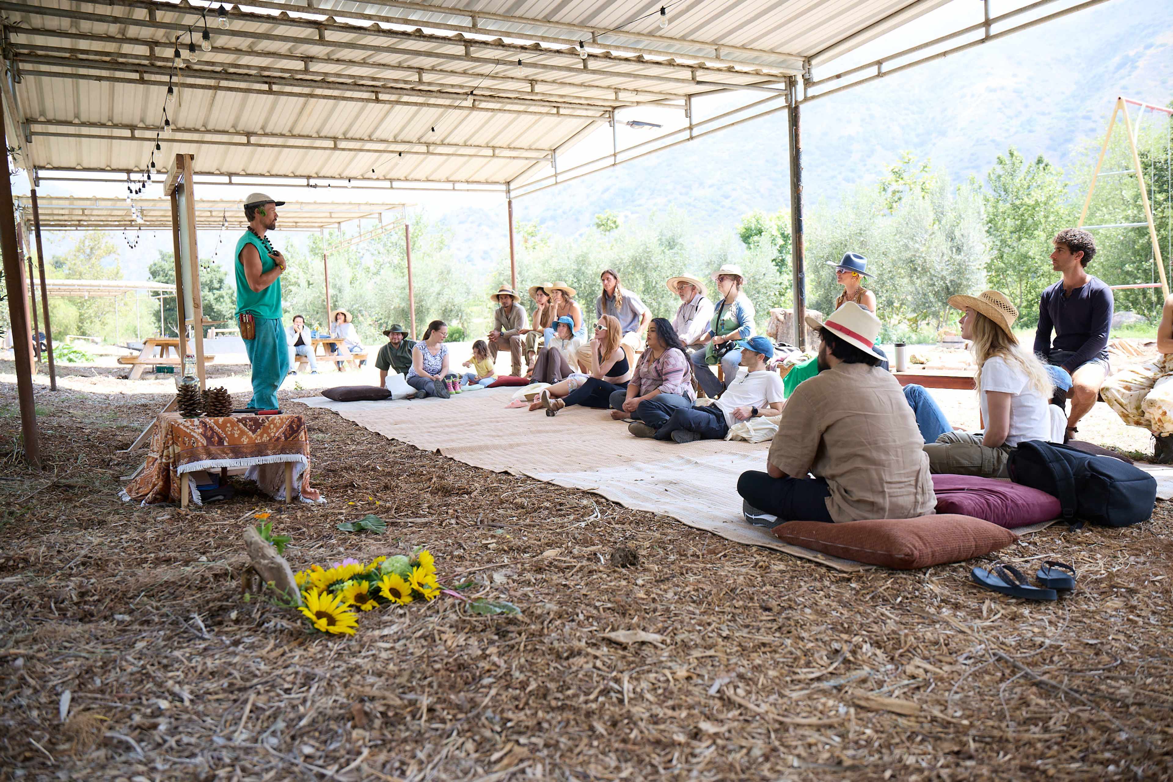 A group of people is seated on the ground in a covered outdoor area, listening to a speaker in front of them, with trees and mountains visible in the background.