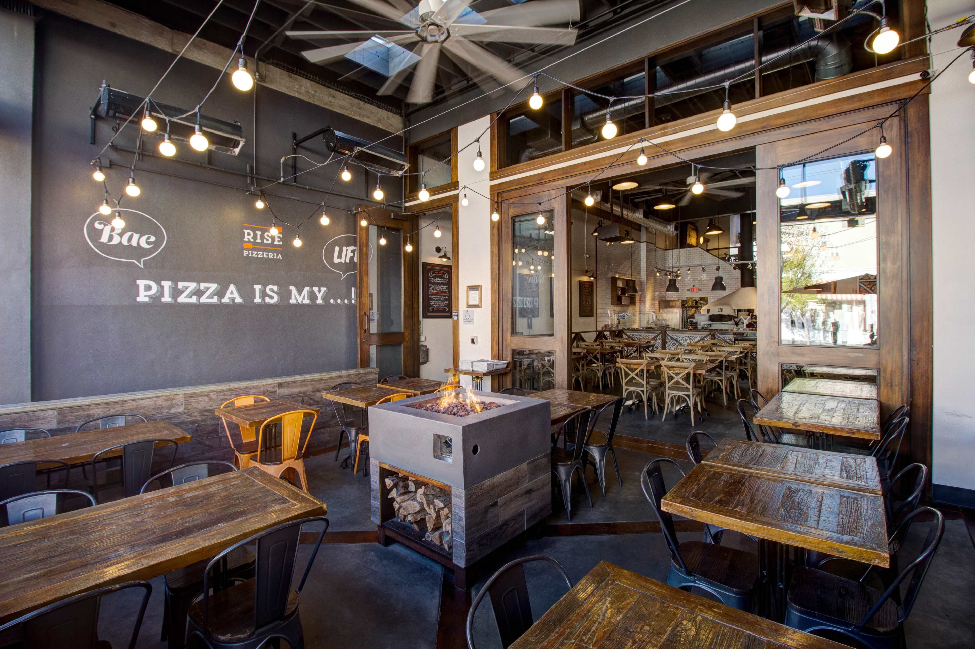 A modern pizza restaurant with wooden tables, metal chairs, and a central fire pit, illuminated by string lights.