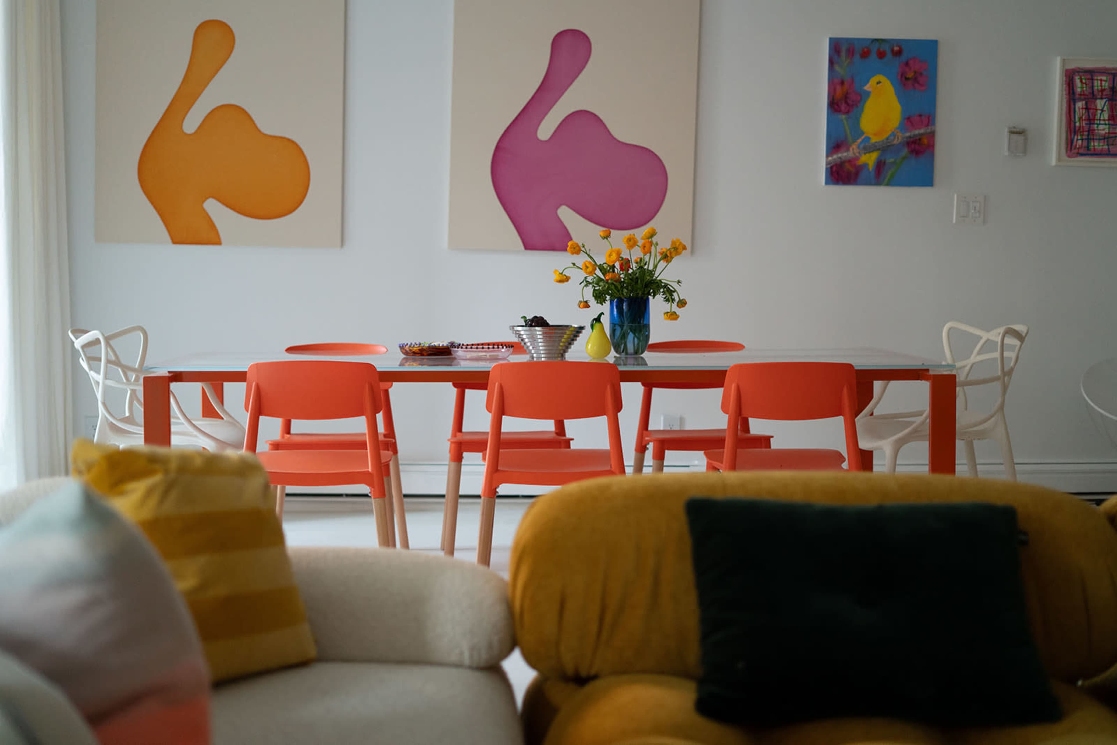 A dining area features a red table surrounded by orange chairs, with two large art pieces on the wall and a vase of flowers at the center.