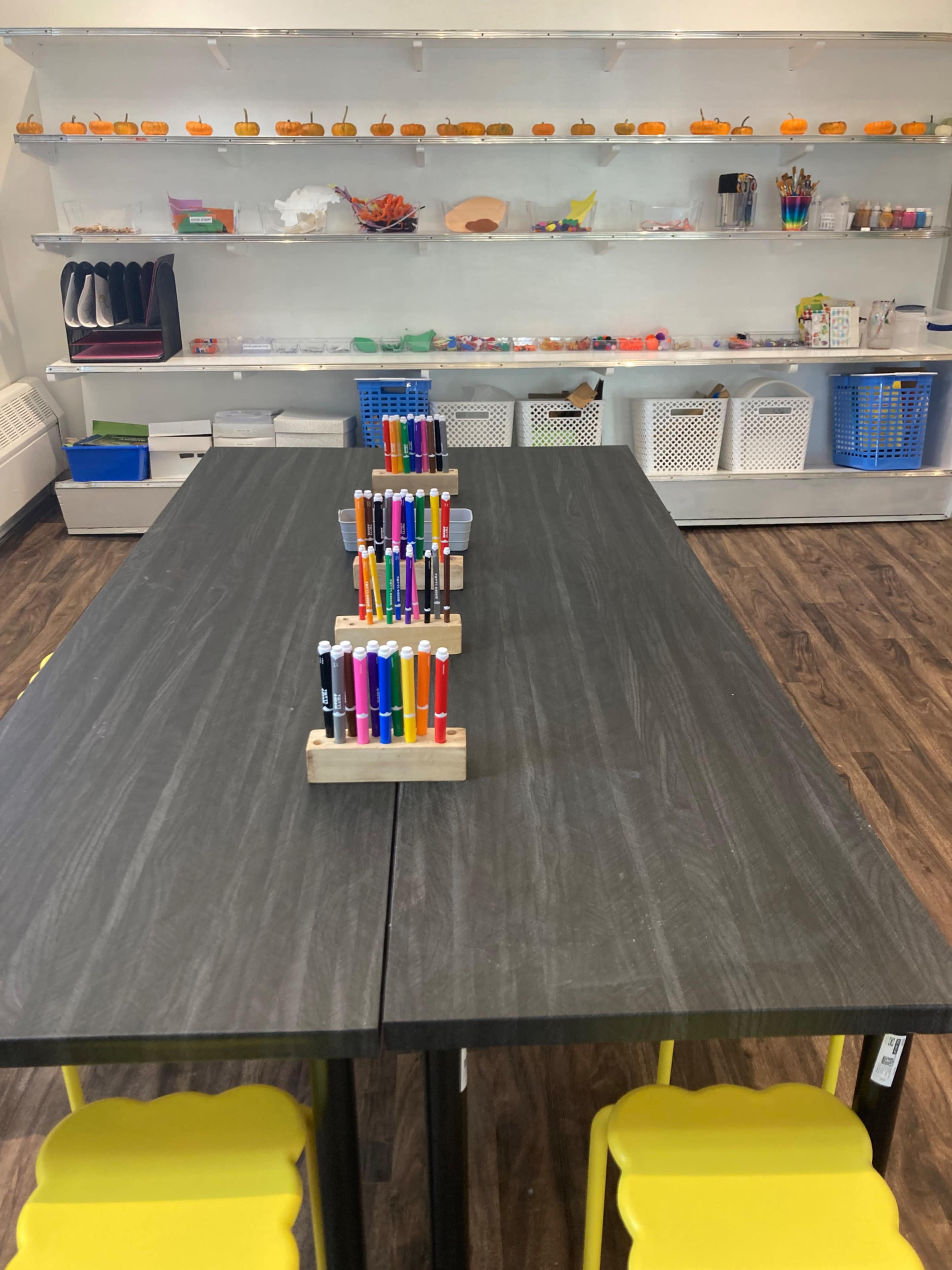 A classroom setup features two large black tables with a holder of colorful markers in the center, and shelves filled with supplies and decorations in the background.