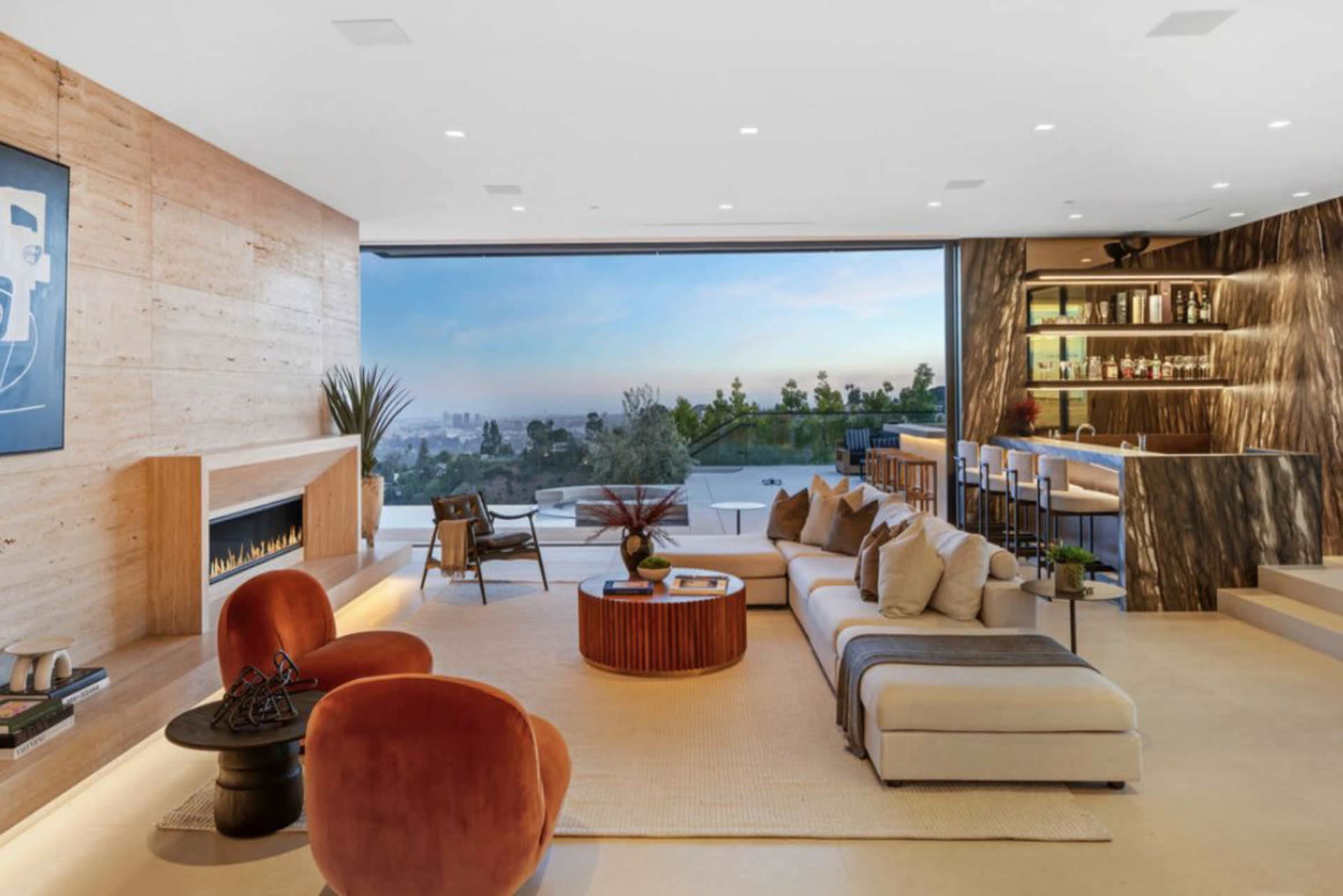 The image depicts a modern living room with large windows offering a view of a city skyline, featuring a sleek design with a fireplace, a sectional sofa, and an open bar area.