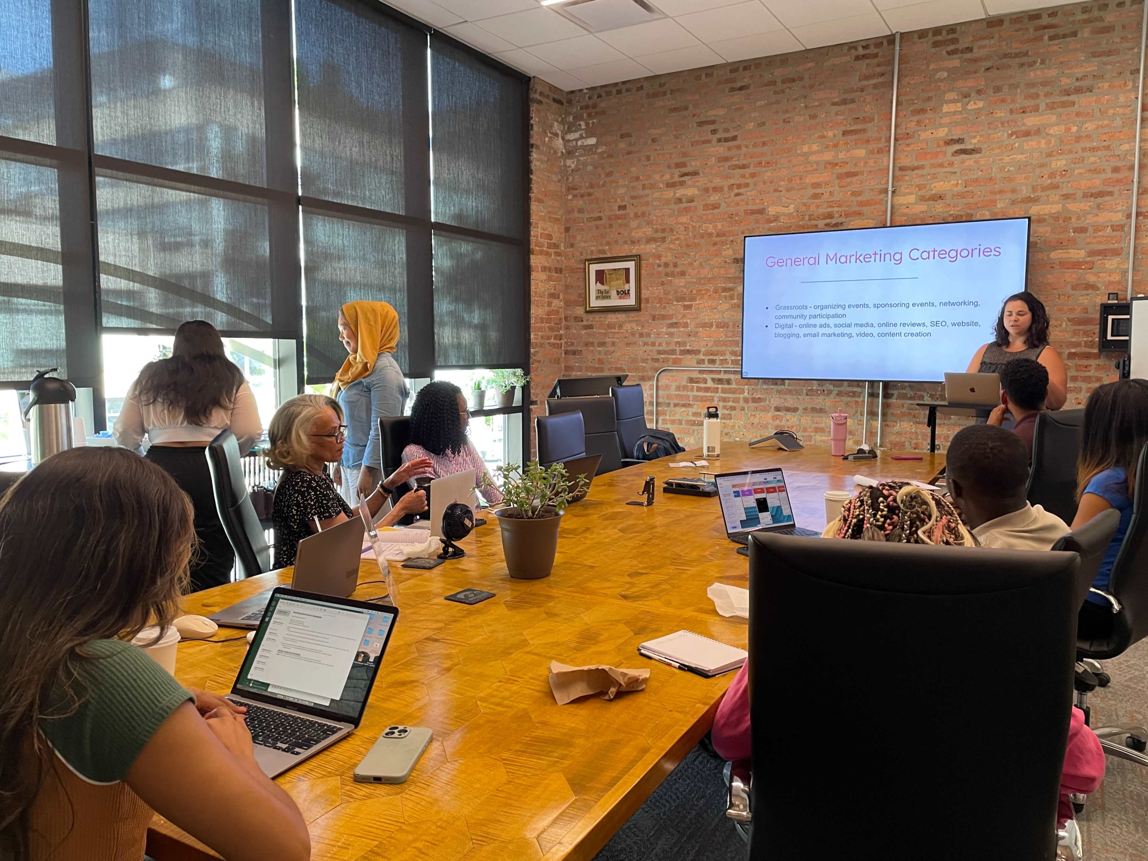 A group of people is gathered around a conference table in a meeting room, where one person is presenting information on marketing categories while others are engaged with laptops and taking notes.