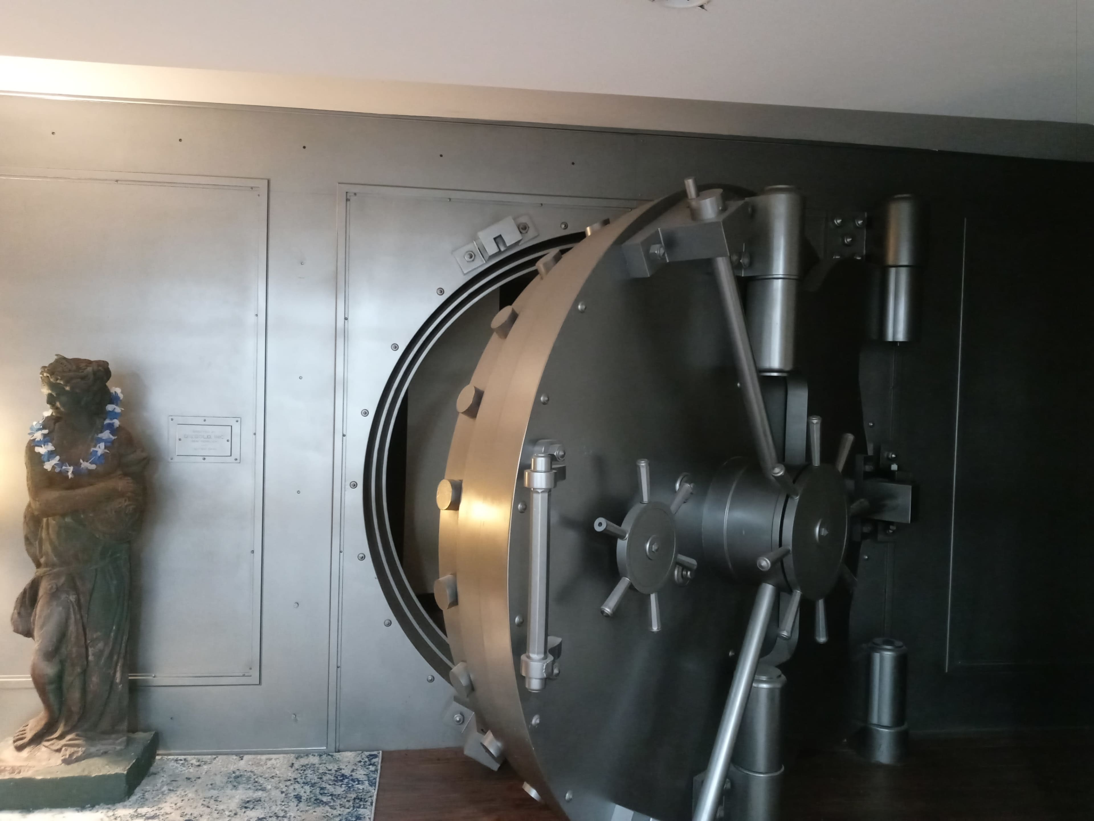A large, metallic safe door is partially open next to a statue covered in a lei.
