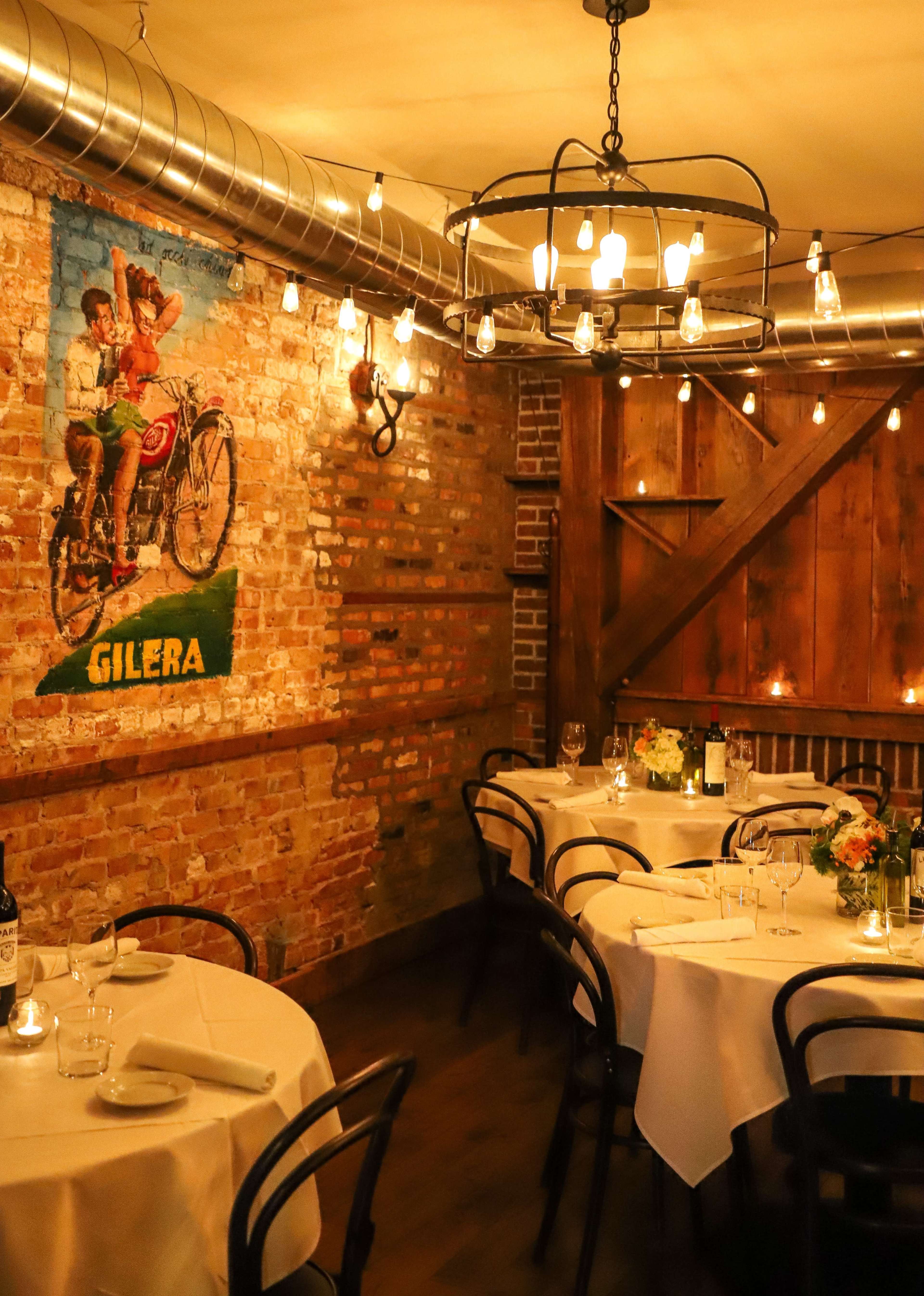 The image shows a warmly lit restaurant interior with wooden beams, brick walls, vintage bicycle art, and elegantly set dining tables.