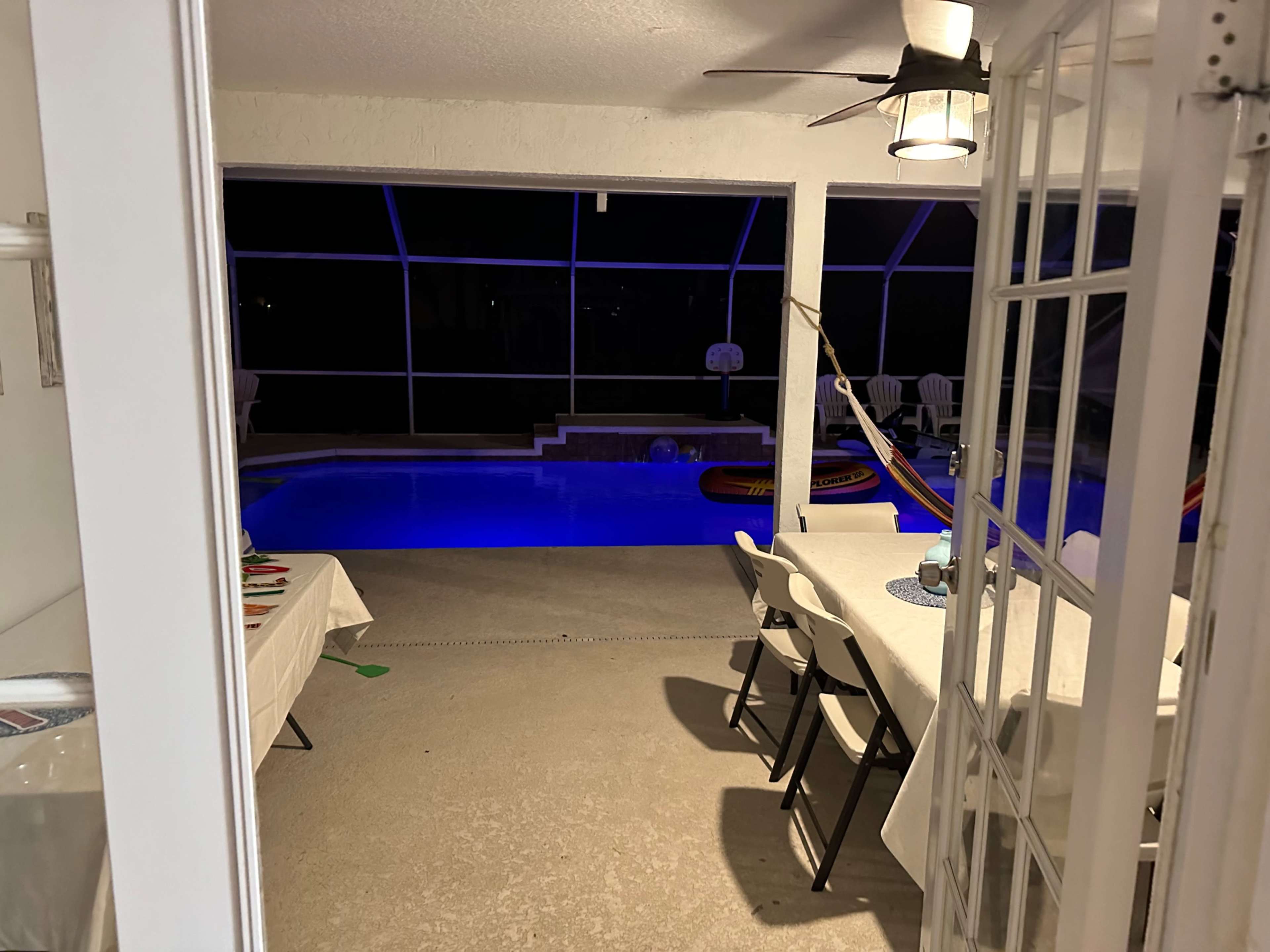 The scene shows a view through a glass door into a well-lit pool area featuring a blue illuminated swimming pool and seating around the table.