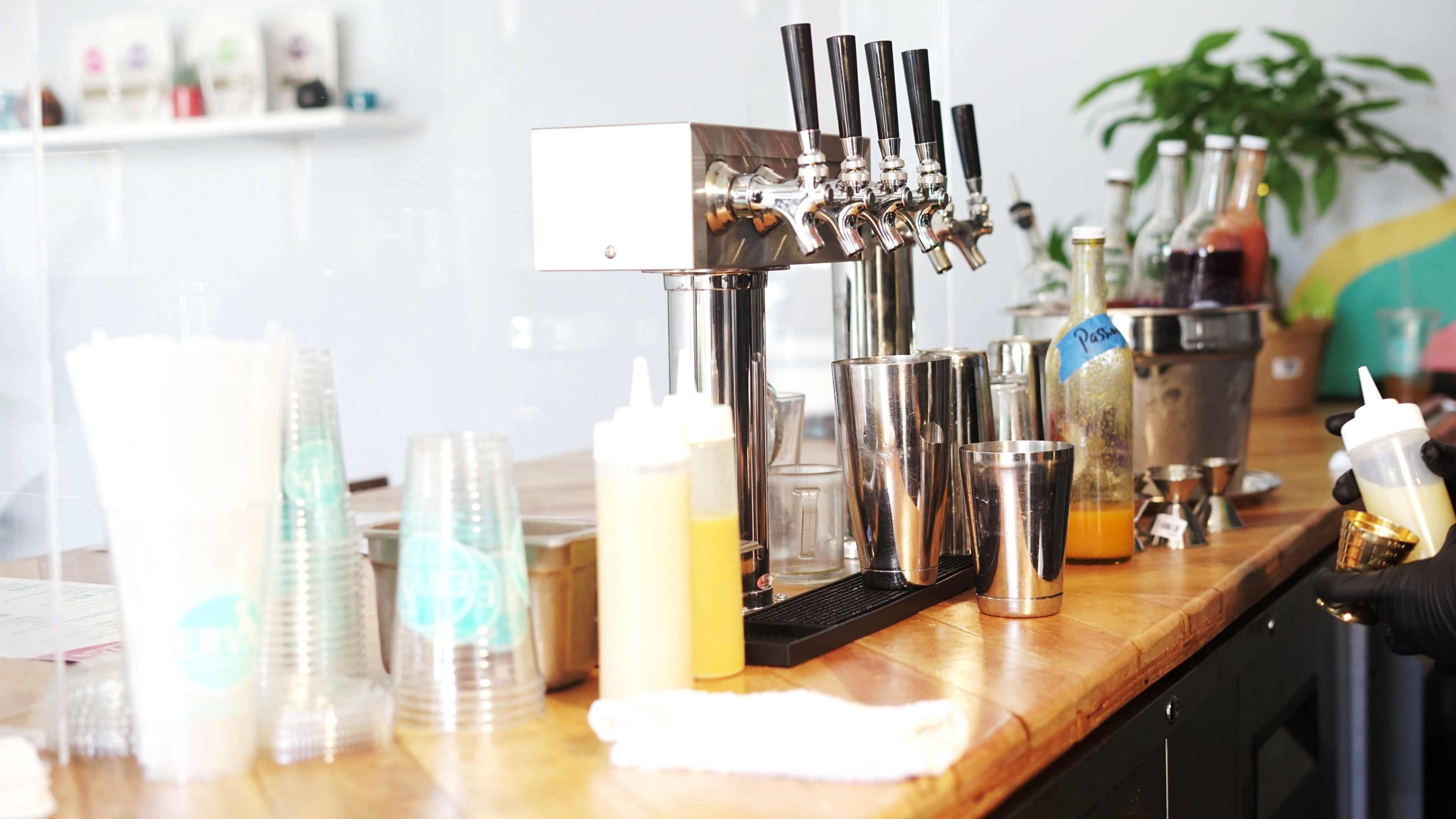 A bar counter with multiple taps for beverages, various containers for condiments, and a plant in the background.