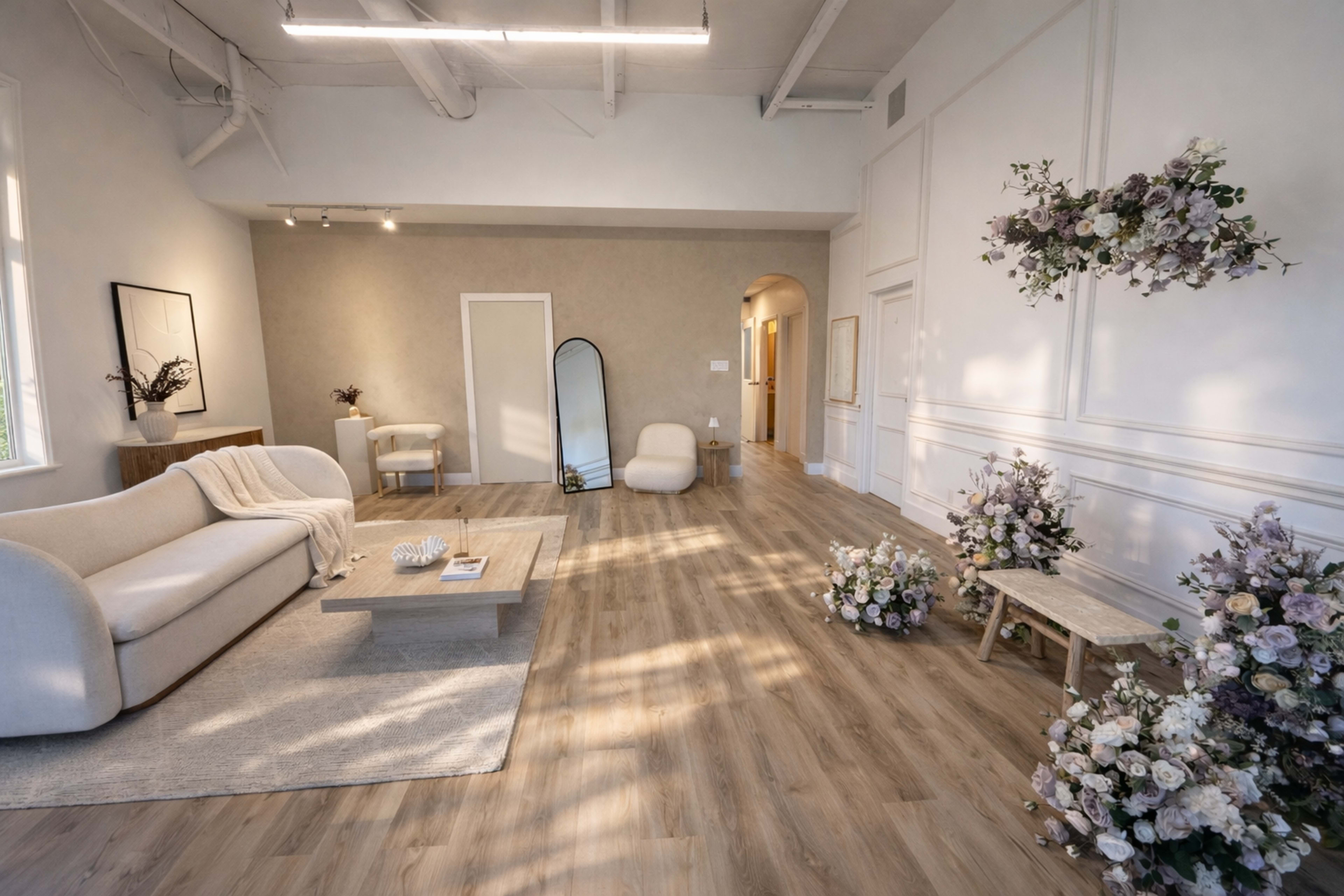 The image shows a spacious, minimalistic living area featuring a light-colored sofa, a wooden coffee table, and floral decorations arranged in the corners.