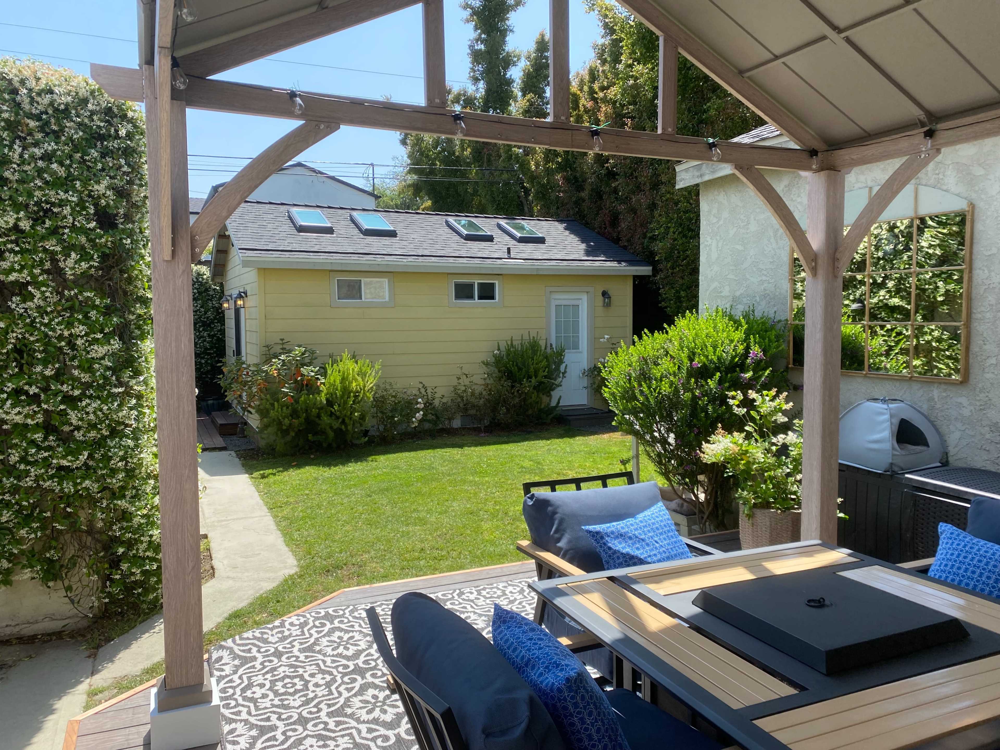 The image shows a backyard area with a wooden pergola, outdoor seating, and a garden leading to a yellow shed.