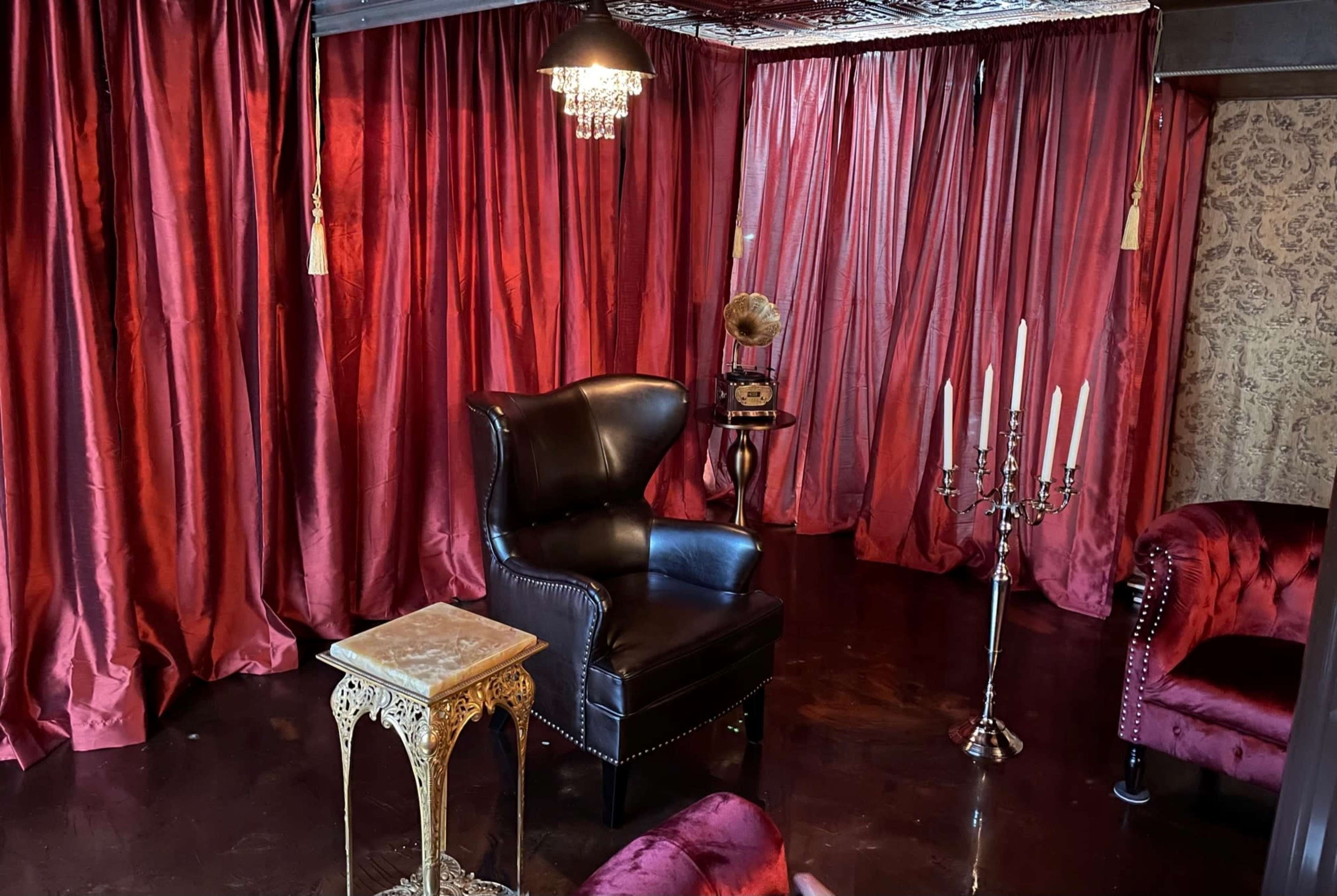 The room features dark red curtains, a black wingback chair, a small vintage table, a candelabra with candles, and a gramophone.