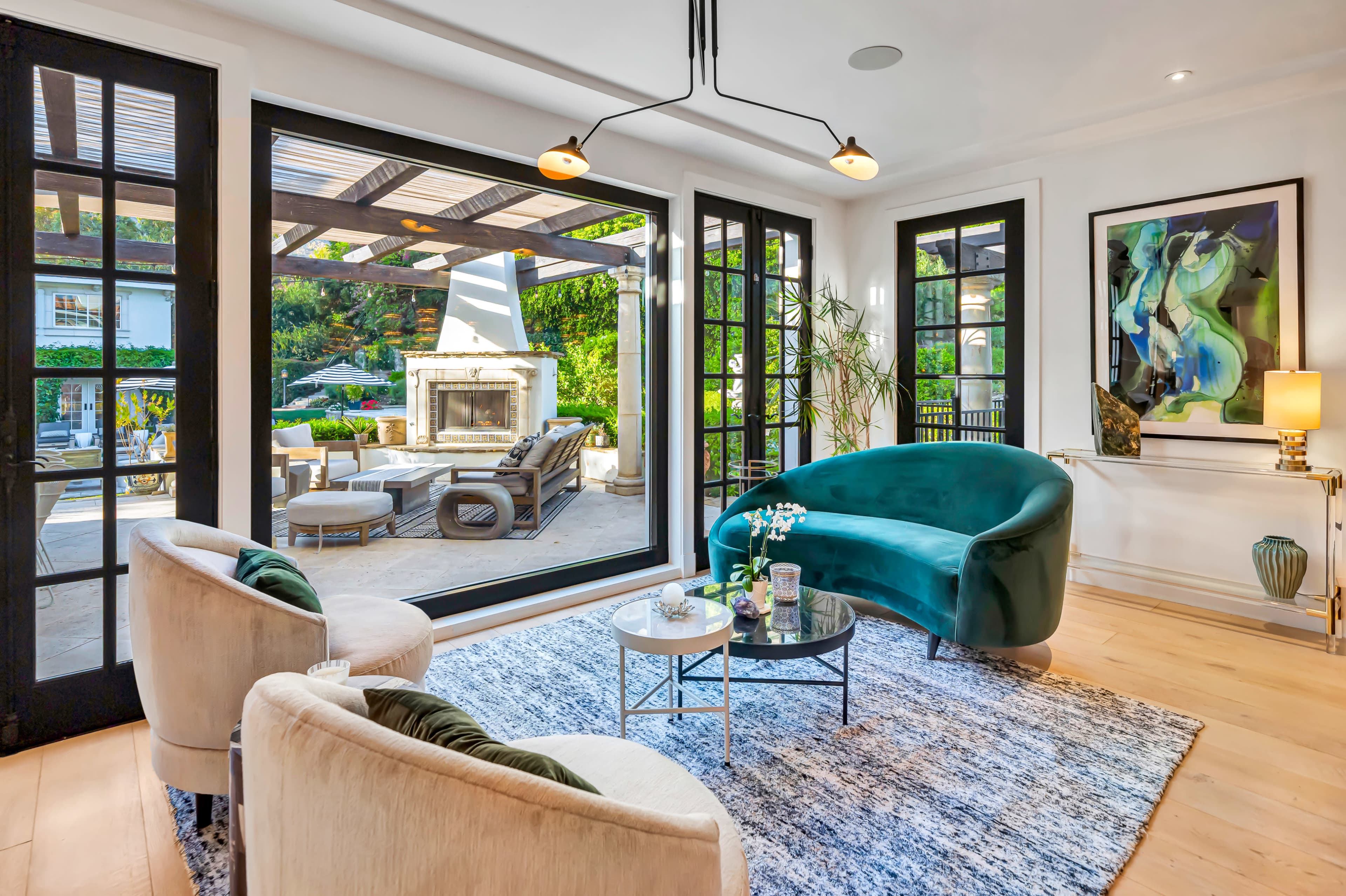 The image shows a modern living room with large windows, featuring a green velvet sofa, two cream-colored chairs, and a coffee table arranged on a textured rug, overlooking an outdoor patio area.