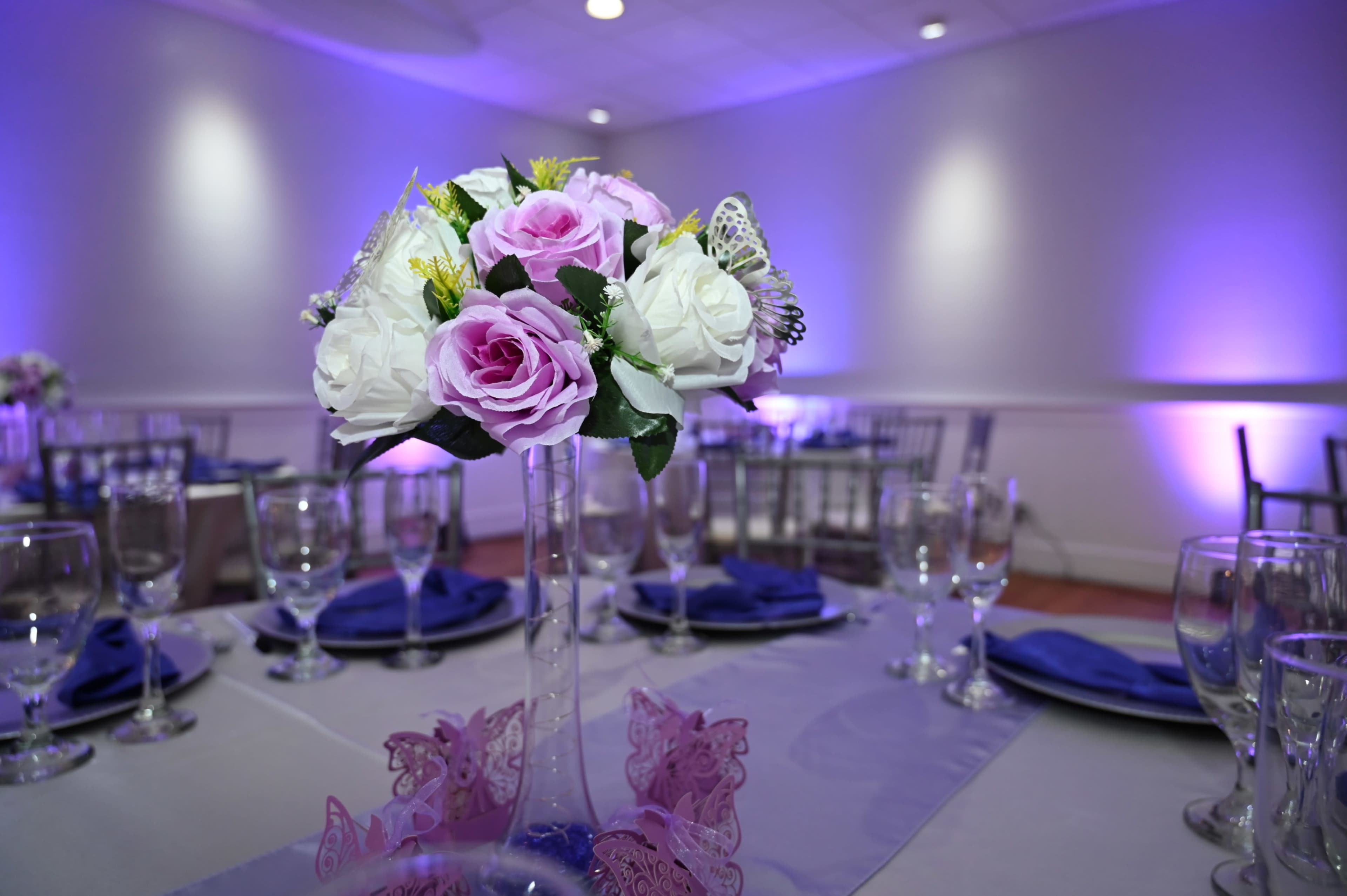 A centerpiece of pink and white roses is displayed on a dining table set with blue napkins and glassware under purple lighting.