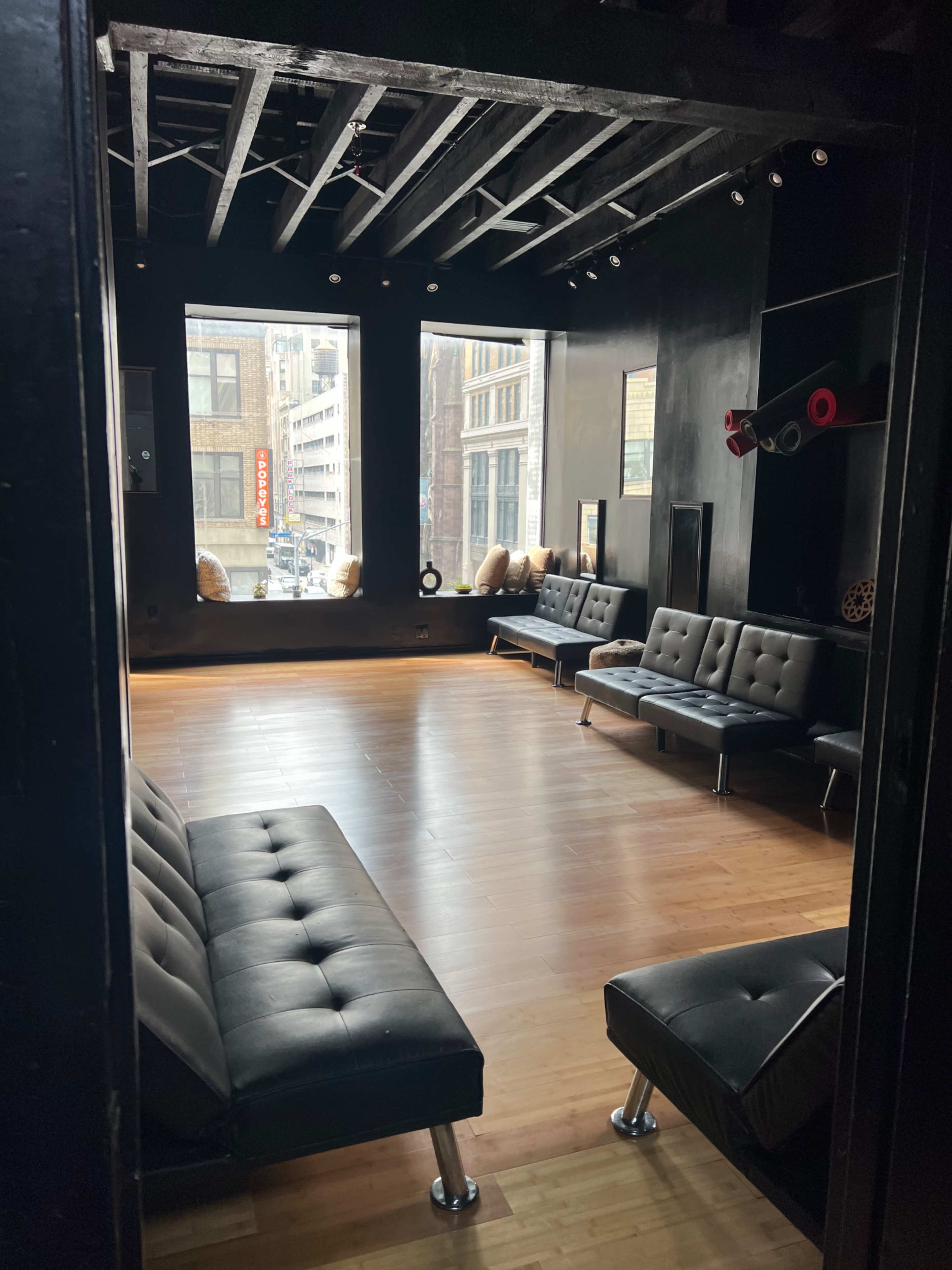 A spacious, minimalist room with black walls, wooden floors, and several black sofas arranged around large windows.