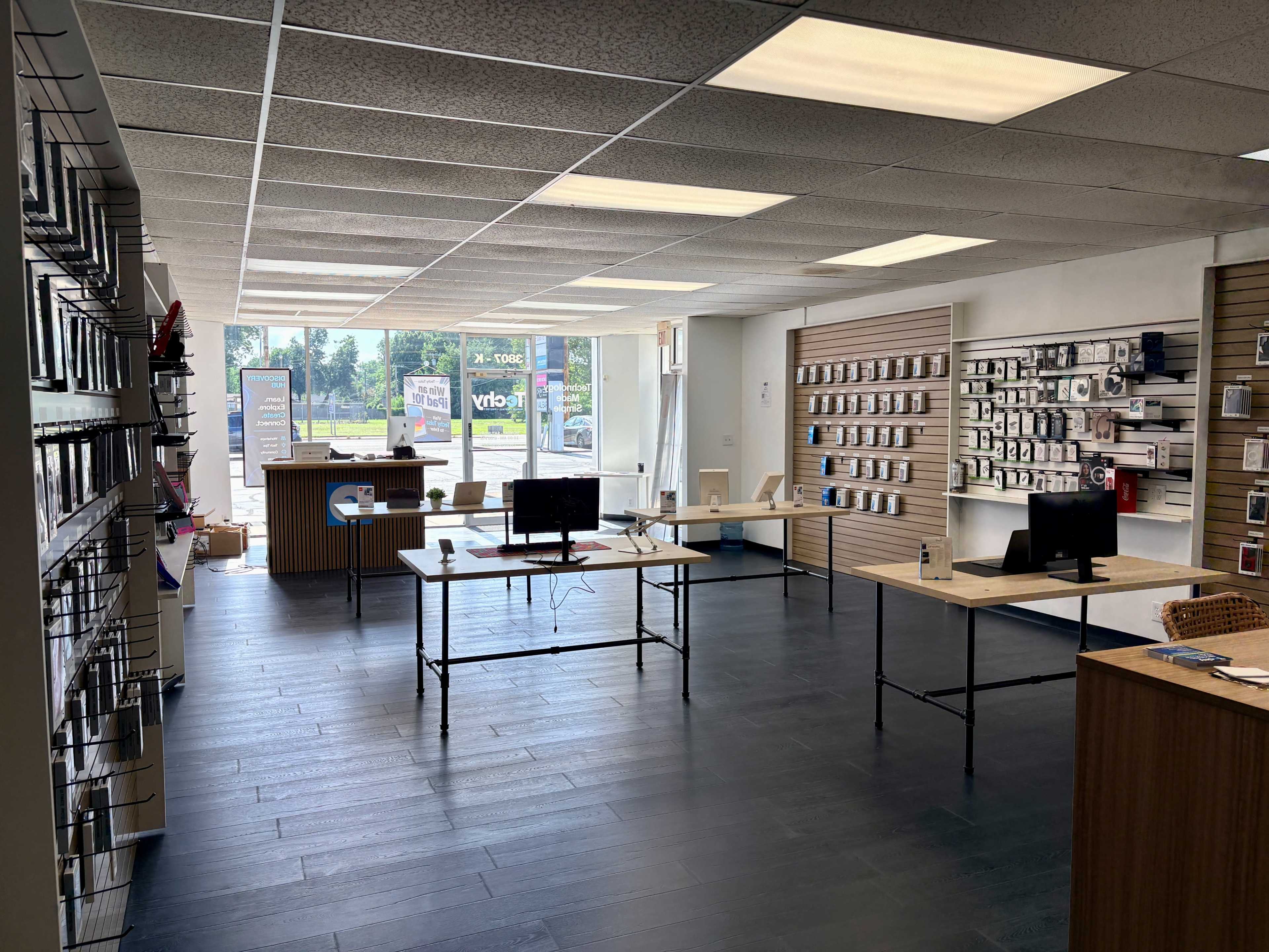 The image shows a modern retail space with several tables displaying electronic devices and accessories, along with shelves lining the walls.
